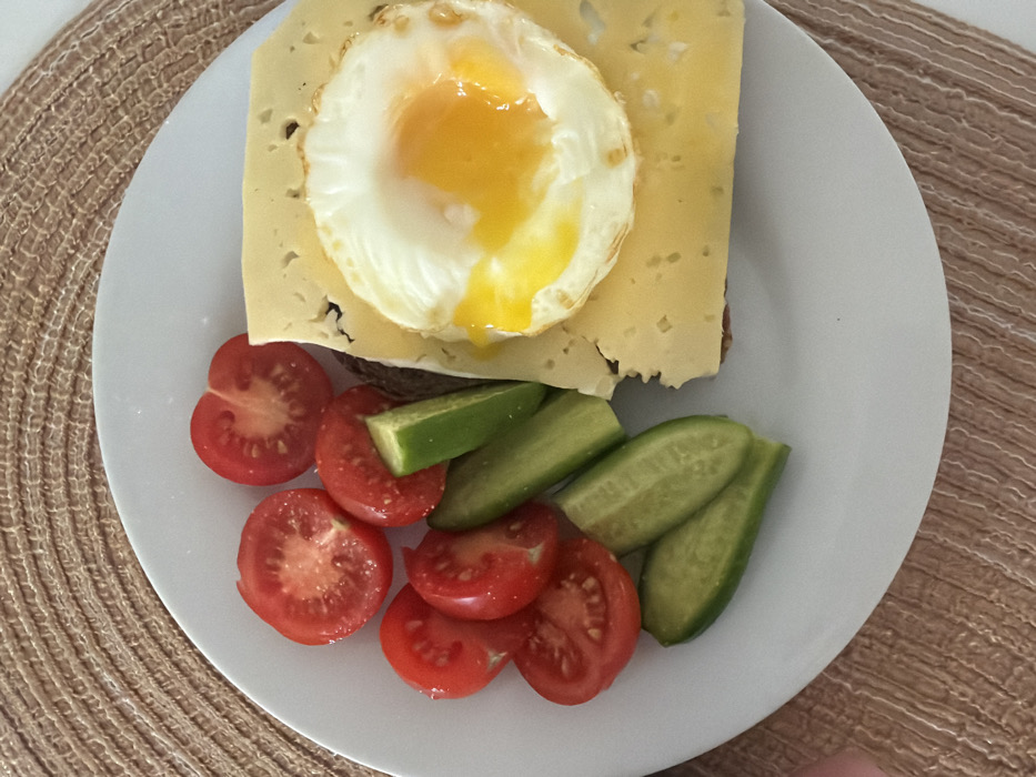 A white circular plate, resting on a beige woven placemat, displays a meal viewed from directly above. On the left side of the plate, there are seven irregular slices of red tomato, some halved, and six elongated green cucumber slices, interspersed among the tomatoes. On the right side, a single, light-yellow rectangular slice of cheese, exhibiting visible holes, partially covers an underlying brown object, possibly bread or toast. Superimposed on the cheese is one fried egg with a solid white albumen and a bright orange-yellow, partially runny yolk. A small amount of yolk has dripped onto the cheese. The image contains no visual information to corroborate the specified location of Tel-Aviv, Israel.