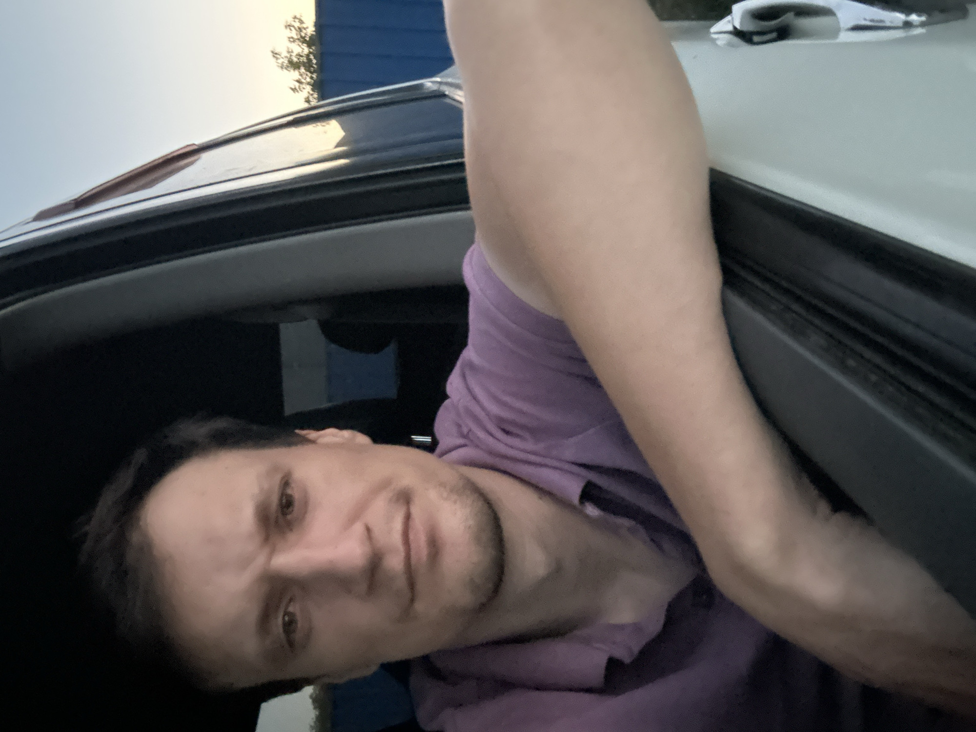 A man, with dark hair and light skin, is leaning out of a car window, with his arm resting on the open door frame. He is wearing a purple polo shirt. The man is looking directly at the camera with a serious expression. The background shows a blue fence and trees under a clear sky, suggesting an outdoor setting, possibly in Shatava, Ukraine. The lighting indicates it is either early morning or late evening.