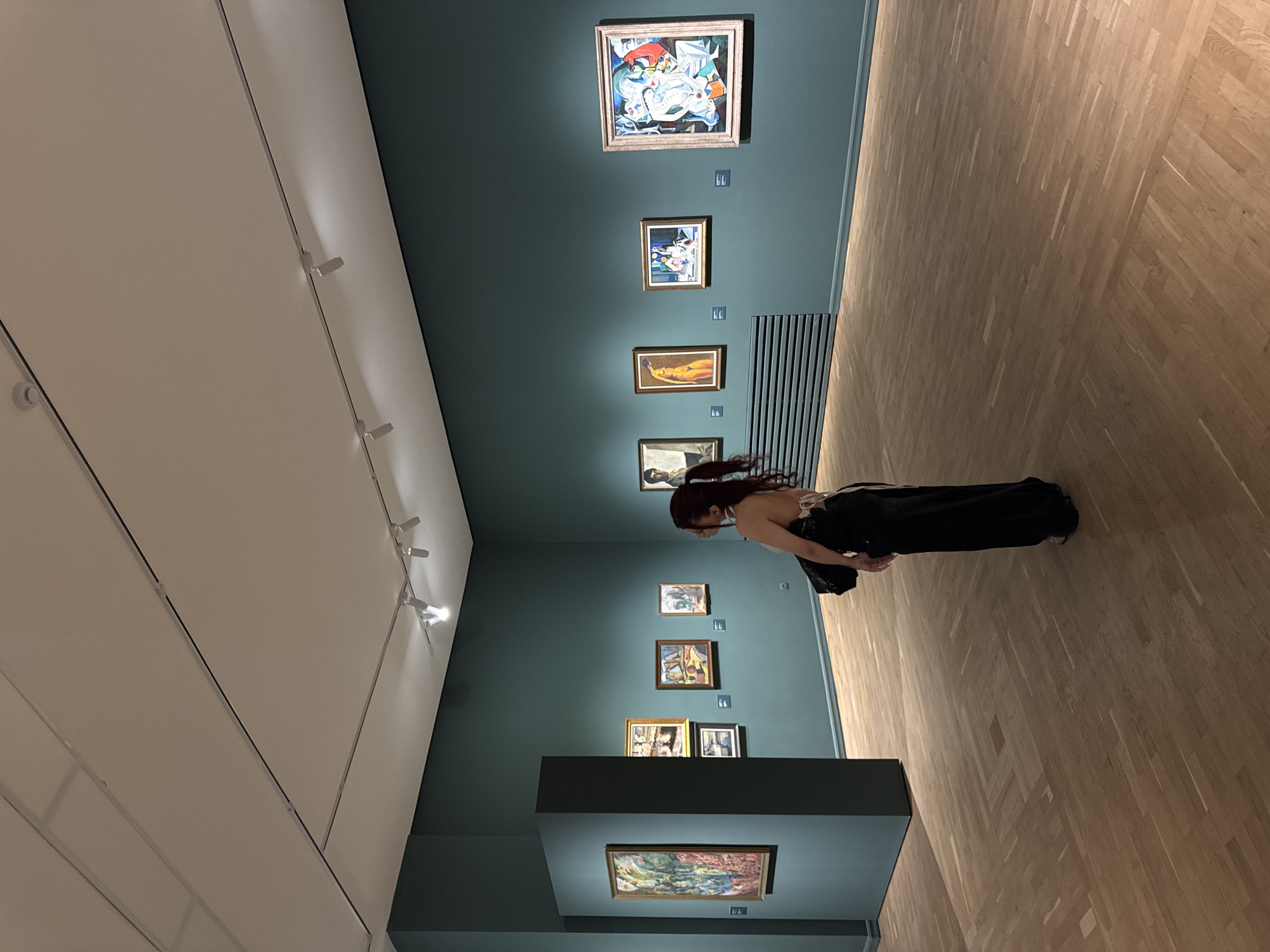 A person with dark hair pulled into a ponytail, wearing a black floor-length dress with a white top and a black purse over their shoulder, stands in a museum gallery, looking at paintings on a teal-colored wall. The gallery has a polished wooden floor with a parquet pattern and is lit by track lighting. Several framed paintings are displayed on the wall, including a large one on the left wall and a row of paintings on the back wall. The setting is an art museum, likely in Yerevan, Armenia, given the user's request.