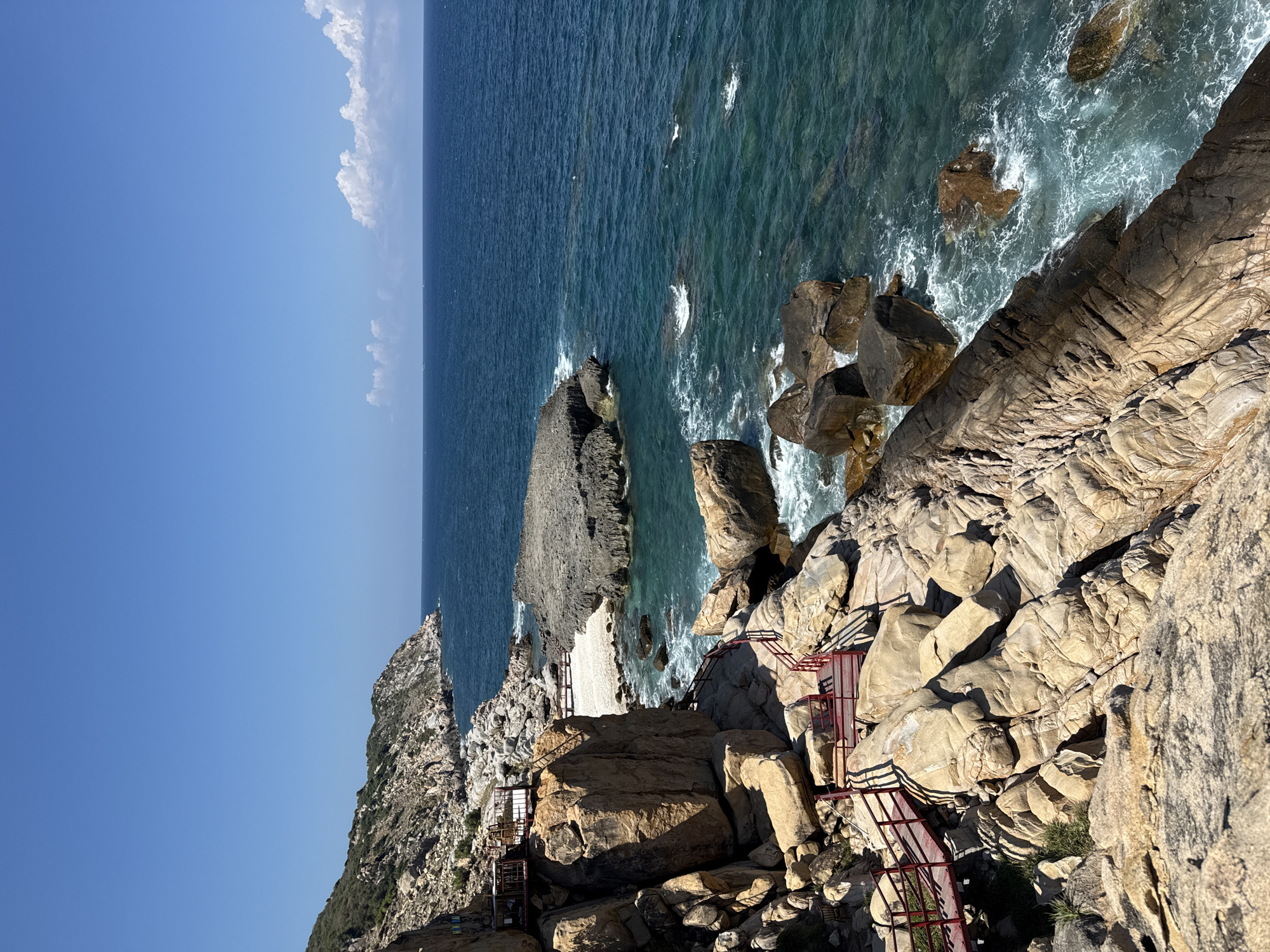 The image depicts a coastal scene in Vinh Hai, Vietnam. A rocky cliff face dominates the foreground, with a constructed metal staircase and viewing platforms built into the rocks. The staircase is painted red. The sea is visible beyond the rocks, with waves crashing against the shore. The sky is blue with some white clouds. There are no people visible in the image.