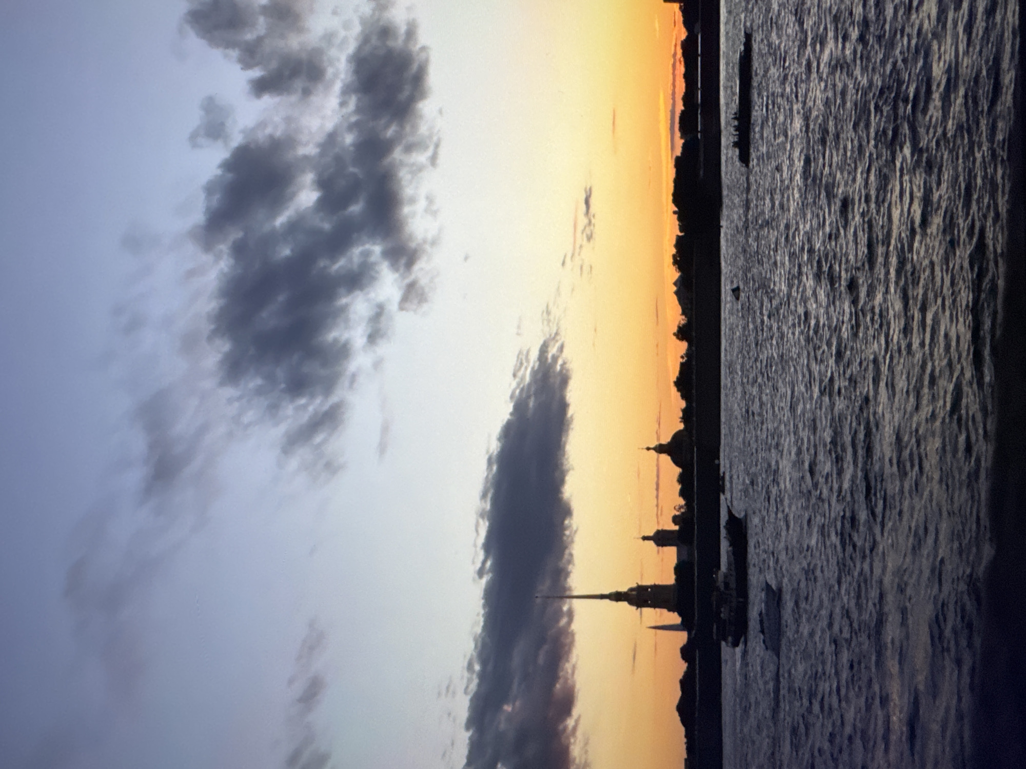 The image depicts a sunset over a body of water, with a silhouetted cityscape in the background. The sky transitions from a pale blue on the left to a vibrant orange and yellow on the right, indicating the sun is setting. Several dark clouds are scattered across the sky, adding texture and depth.
The cityscape features several distinctive architectural structures, including spires and domes, suggesting a historical or significant urban area. These buildings are entirely in silhouette against the bright sky. The foreground is dominated by the dark, rippling surface of the water, which reflects the colors of the sunset faintly. A small boat is visible on the water, adding a sense of scale to the scene.
The overall mood of the image is serene and picturesque, capturing the beauty of twilight. The lighting suggests it is late evening. No people are clearly visible, but the presence of the cityscape and boat implies human activity in the area. There is no visible text in the image. The composition, with the horizon line separating the sky, cityscape, and water, creates a balanced and visually appealing scene.