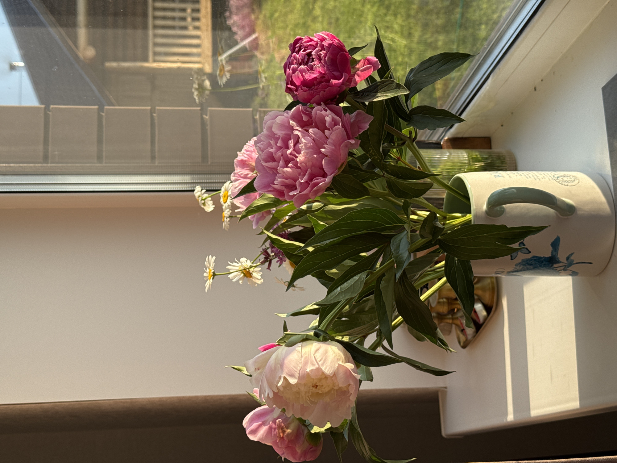 A close-up, eye-level, horizontal shot captures a bouquet of pink peonies and white daisies in a white ceramic vase with blue floral patterns and a light green handle. The vase sits on a white windowsill, casting a shadow to the left, suggesting the sun is positioned to the right. The background shows a window with a view of a wooden house and a manicured lawn, bathed in warm, late afternoon sunlight. The setting is likely a residential area in Izhevsk, Russia, given the architectural style of the house and the commonality of peonies in Russian gardens during summer. No people are visible in the image.