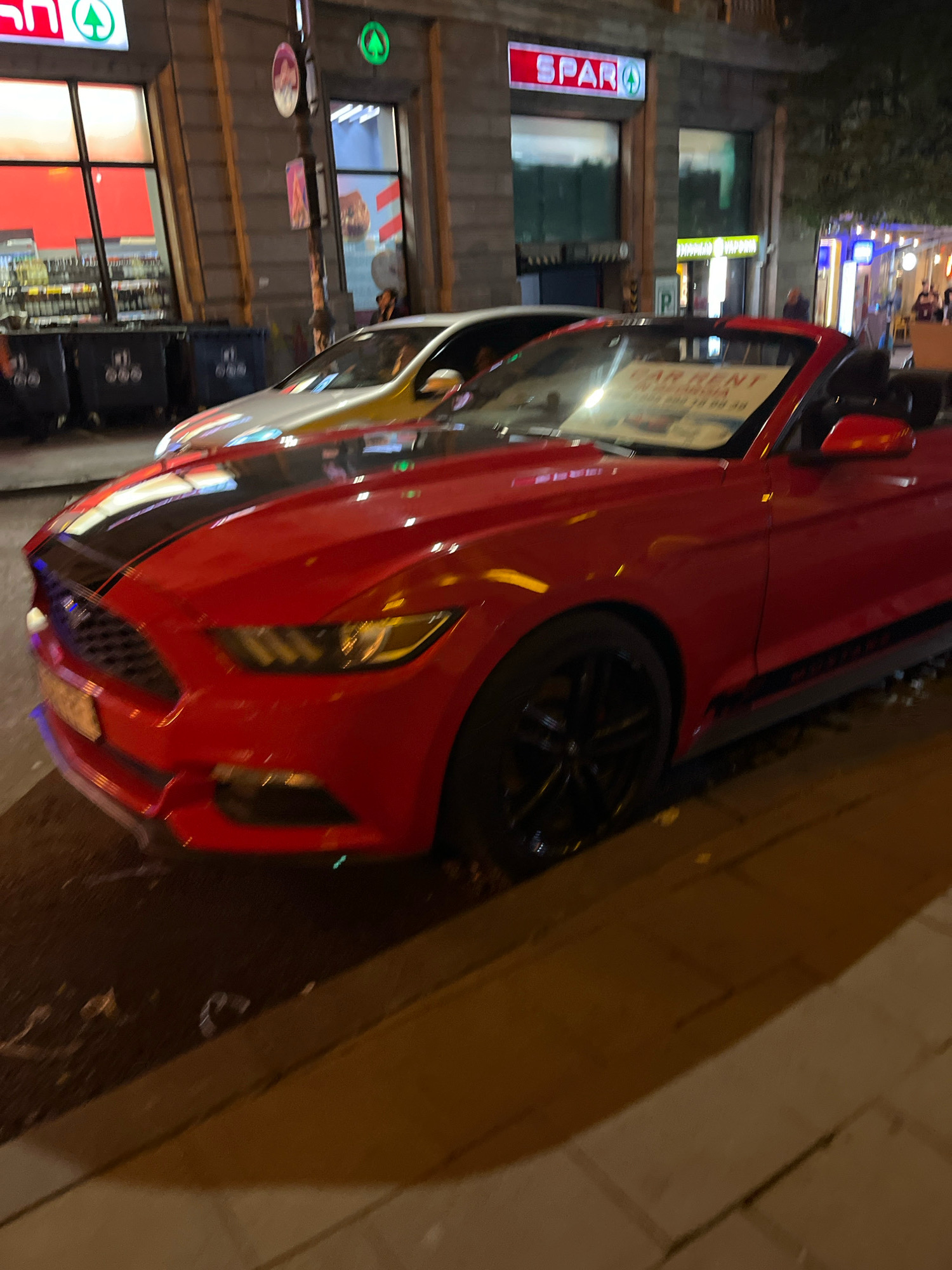 A red Ford Mustang convertible is parked on a city street at night, with a "CAR RENT" sign on its windshield. The car is angled towards the viewer, with its front left side and hood most prominent. A white car is parked behind it. In the background, a SPAR supermarket is visible, along with other storefronts and streetlights. The scene appears to be in Tbilisi, Georgia, based on the visual cues. The overall lighting suggests it is nighttime.