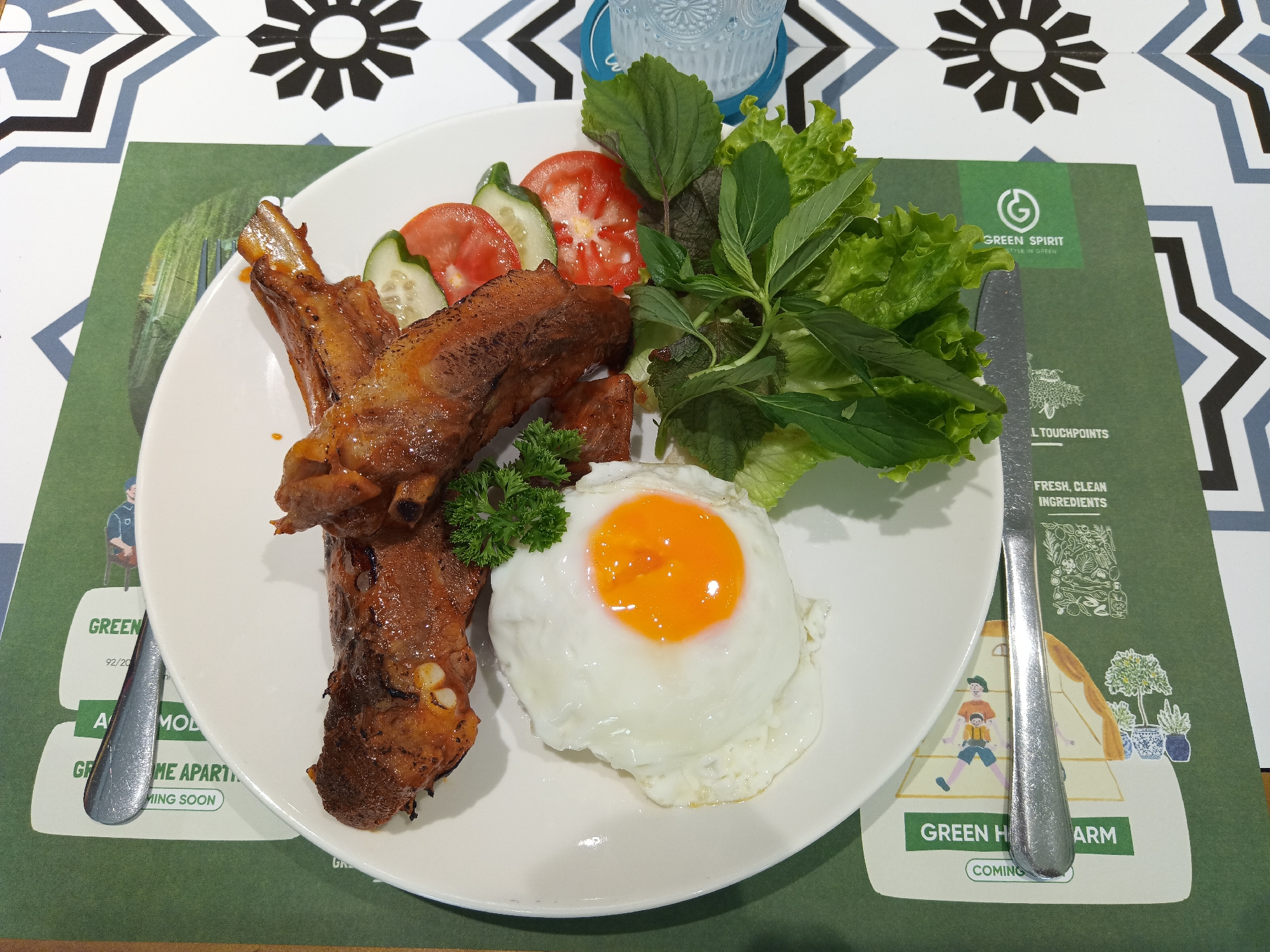 A plate of food sits atop a green and white decorative placemat in Nha Trang Ward, Vietnam. The dish consists of glazed ribs, a sunny-side-up egg, tomato and cucumber slices, and various greens including lettuce, basil, and parsley. A silver knife is positioned to the right of the plate, resting on the placemat. The placemat itself has a design with text reading "Green Spirit, Style in Green" and other related information, suggesting an environmentally conscious theme. Above the placemat, part of a patterned table or countertop is visible, consisting of a design of black and gray geometric shapes on a white background. A partially visible glass item sits above the plate.