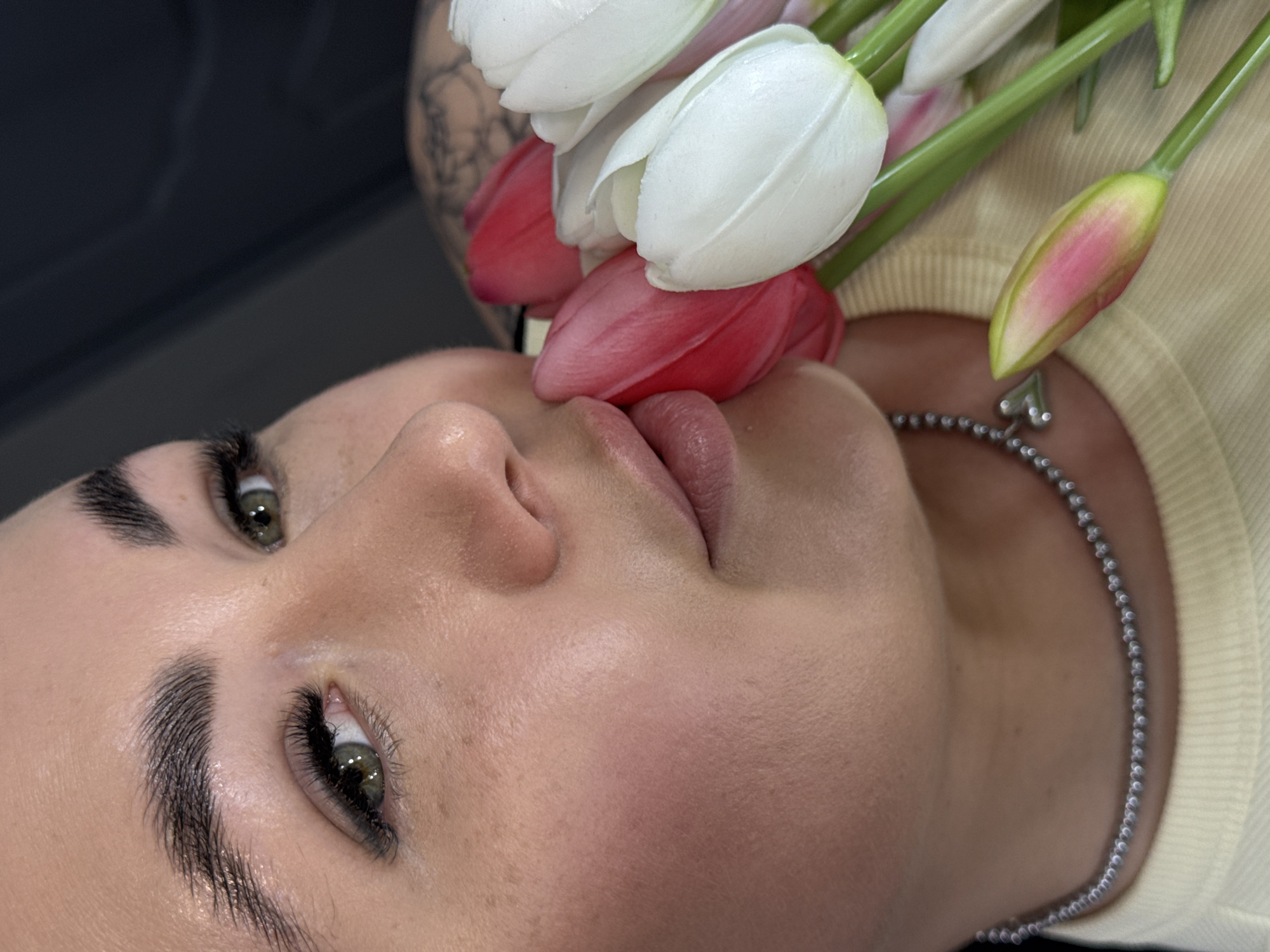 A close-up, angled shot depicts a person's face positioned amongst white and pink tulips. The individual has fair skin, dark eyebrows, and green eyes with long lashes. A silver beaded necklace with a heart-shaped pendant is visible around their neck. They are wearing a light yellow shirt. Part of a tattoo is visible in the background. The scene is likely set indoors. The location is the city of Chaikovsky, Russia.