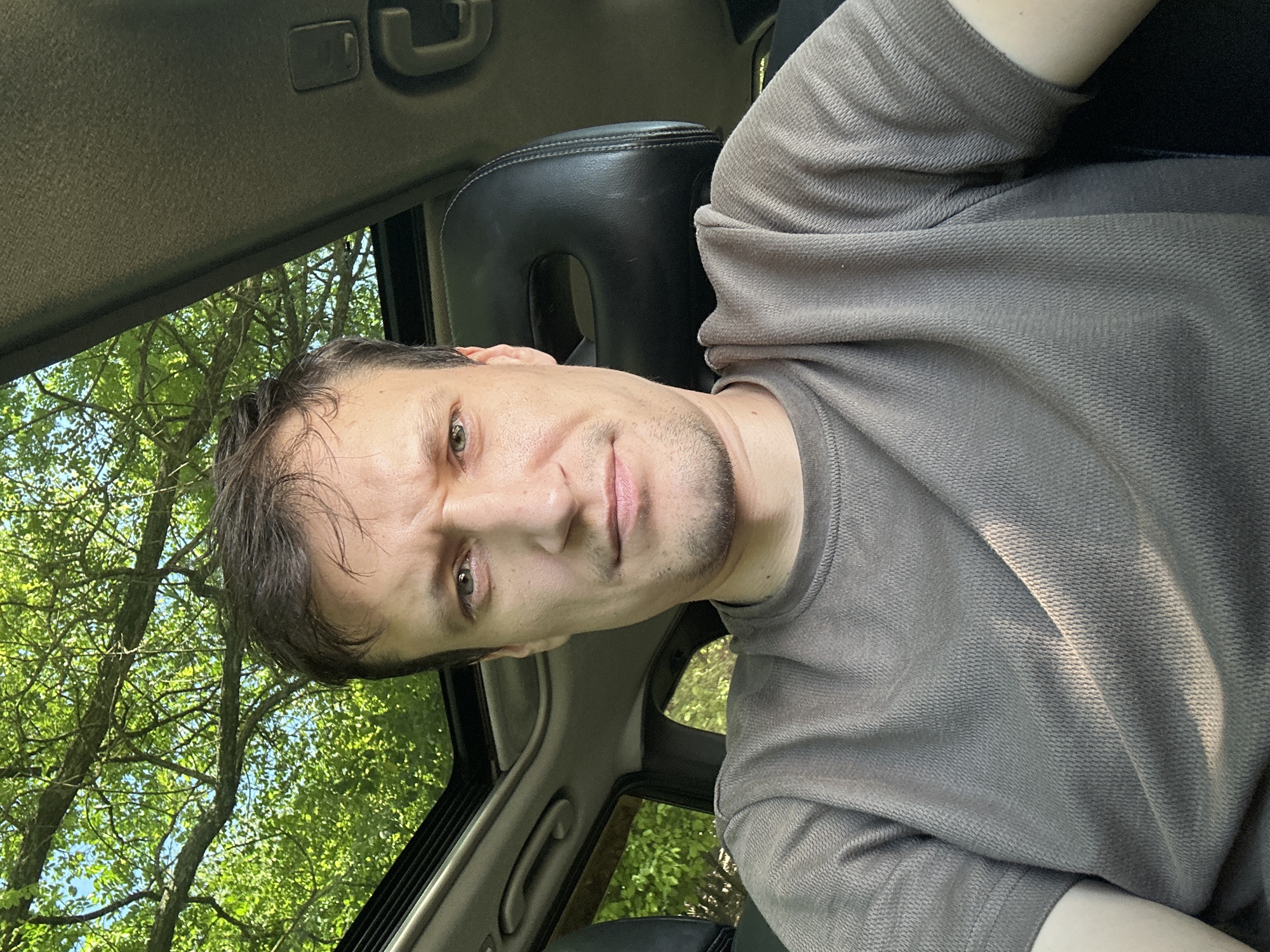 A man is taking a selfie inside a car with a sunroof open. Sunlight filters through the trees and the sunroof, casting shadows on his face and brown, textured t-shirt. The background shows green foliage and the car's interior, including a headrest. The man's expression is a slight frown. The location is not specified in the image or provided context.