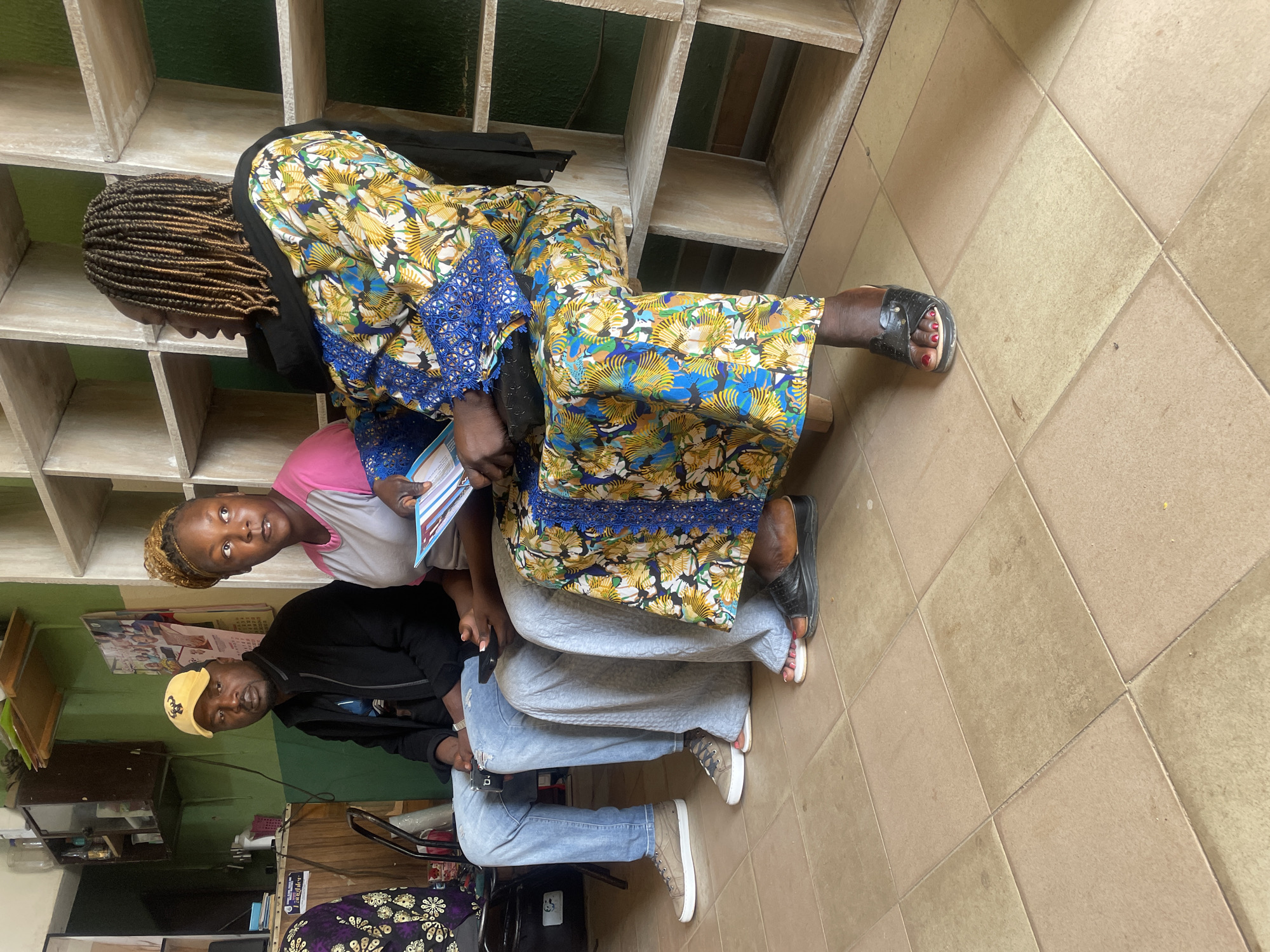 In an indoor setting in Jos, Nigeria, three individuals are seated on a bench against a backdrop of shelves. A man in a yellow cap and black jacket sits on the left, wearing jeans and brown sneakers. Next to him, a woman in a pink and grey shirt is seated. To the right, another woman, wearing a colorful patterned dress with blue trim and a black head covering, is seated. She is holding a pamphlet or booklet. The floor is tiled in a beige, square pattern. The shelves behind the individuals appear to be made of light-colored wood. The overall impression is of a waiting area or a communal space.