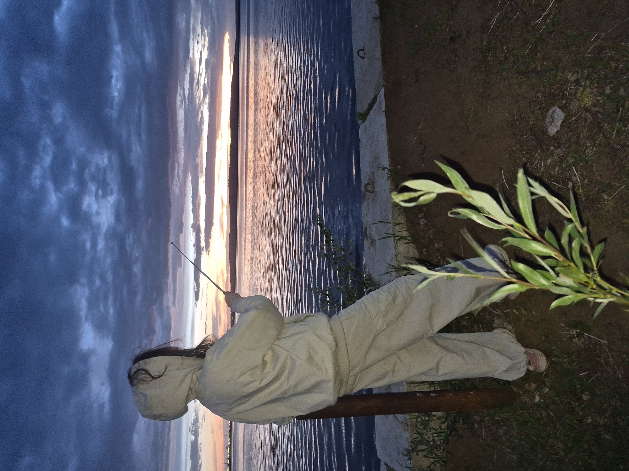 A sideways medium-shot photograph captures a person wearing a light beige hooded garment standing by the river, holding what appears to be a fishing rod. The water is reflecting the colors of the sunset, which blends from blue to orange above it. The person is standing near the edge of the river bank with vegetation and a branch visible next to them. The location is likely in Chaikovsky, Russia.