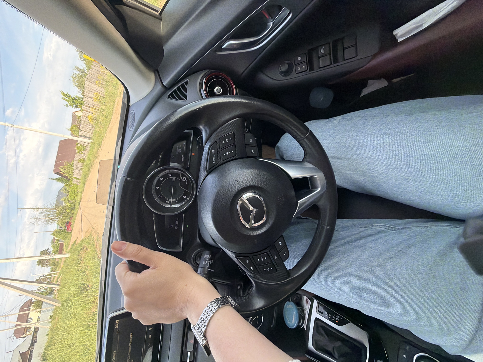 The image shows the interior of a car, specifically a Mazda, with the driver's perspective visible. The steering wheel, dashboard, and part of the driver's hand wearing a silver watch are in the foreground. Through the windshield, a rural or suburban street with houses, trees, and utility poles is visible. The infotainment screen displays Russian text and shows a time of 08:49. Based on this information, and the OCR of "Павел Радонцев", the location is most likely in or near Chaikovsky, Russia. The driver is holding the steering wheel, suggesting they are driving or about to drive.