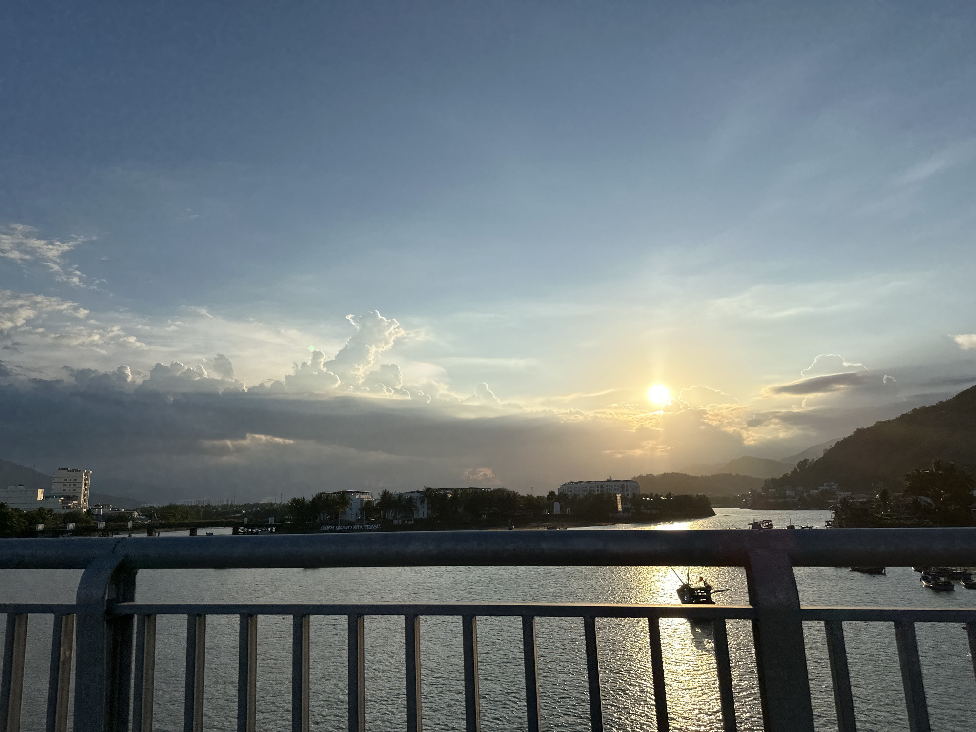 A wide-angle, eye-level shot captures a serene sunset over a body of water in Nha Trang, Vietnam. The sky is a gradient of soft blues and whites, with dramatic, illuminated clouds forming a backdrop to the setting sun. The sun, a bright, diffused orb, casts a golden glow on the water's surface, creating shimmering reflections.
In the middle ground, a low-lying coastline is dotted with buildings and lush green trees. Several boats, some small and traditional, are anchored or gently floating on the water, adding a sense of activity to the tranquil scene. A prominent bridge railing, constructed from dark metal bars, runs across the foreground, framing the view of the water and distant landscape. The water itself appears calm, with subtle ripples reflecting the ambient light. The overall impression is one of peaceful twilight in a coastal city.