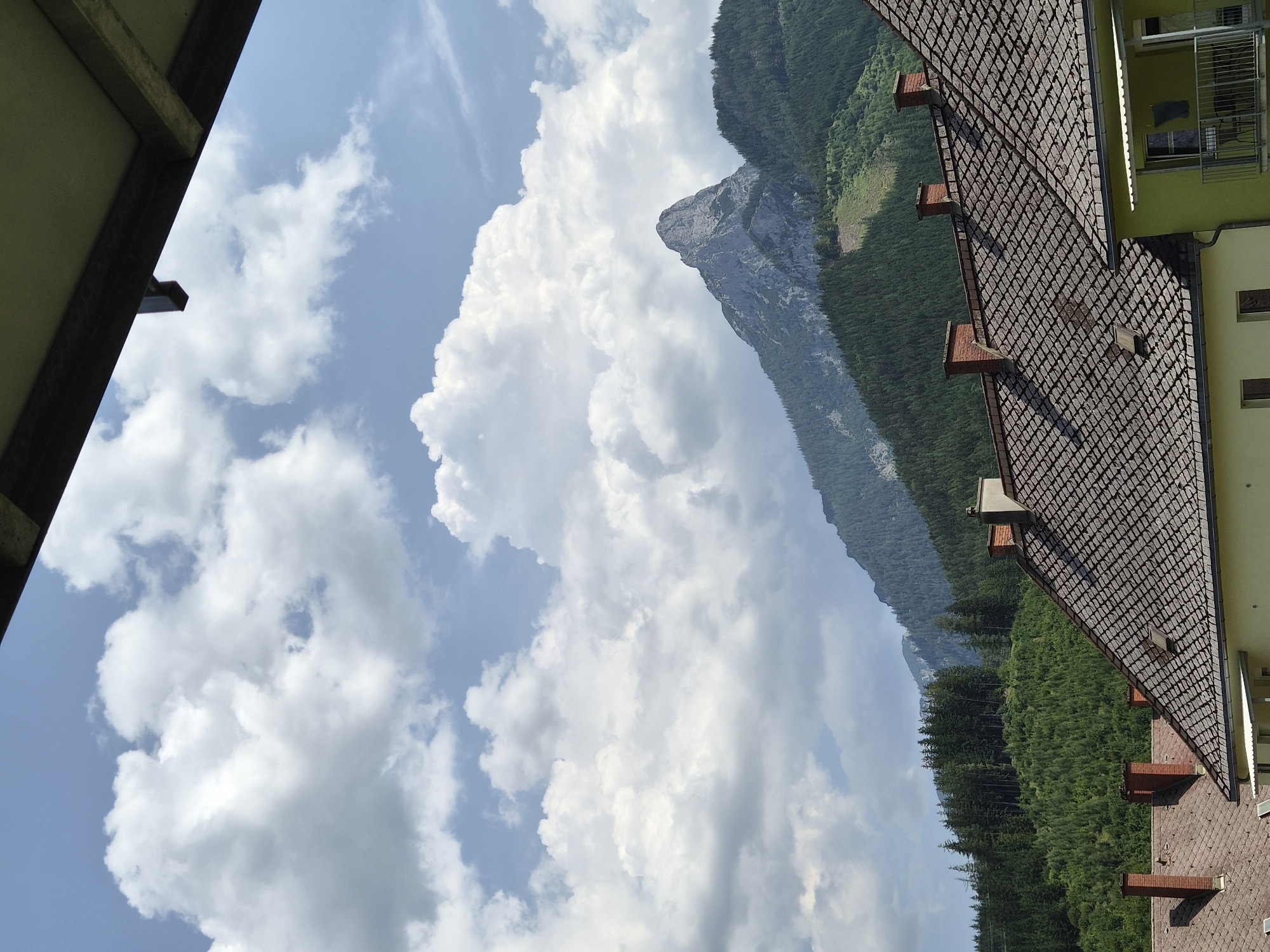 The image, taken from a slightly elevated perspective, captures a mountainous landscape under a partly cloudy sky. In the foreground, the tiled roofs of several buildings are visible, with prominent brick chimneys. The buildings appear to be residential, with one house painted a pale yellow and another a lighter color. Beyond the houses, a dense forest covers the lower slopes of the mountains.
The dominant feature of the scene is a sharp, jagged mountain peak that rises majestically from the forested hills. The peak itself is rocky and appears steep, with visible geological formations. The surrounding mountains are covered in dark green coniferous trees, creating a textured, undulating backdrop. The sky above is a vibrant blue, punctuated by large, fluffy white cumulus clouds that cast shadows on the mountains.
This image likely depicts a view from the city of Eisenerz, Austria, a town known for its dramatic mountain scenery and rich mining history. The juxtaposition of the man-made structures in the foreground with the imposing natural beauty of the mountains suggests a harmonious integration of human habitation with the alpine environment. The lighting indicates it is daytime, possibly late morning or early afternoon, given the brightness and the clear visibility of the details. There are no people or animals visible in the frame. The overall impression is one of a serene and picturesque alpine setting.