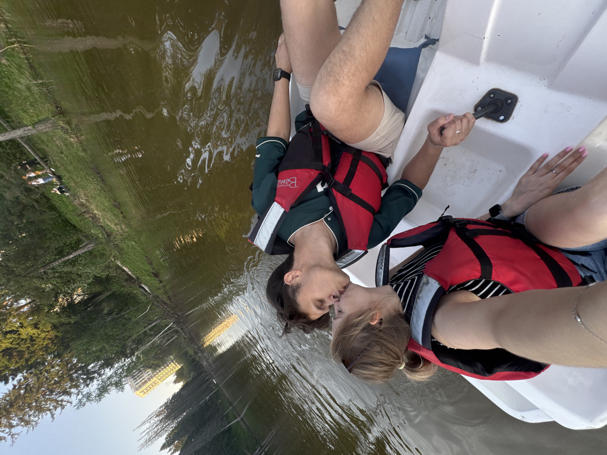 In Izhevsk, Russia, a couple is kissing on a white boat in the water. The man is wearing a dark green jacket with white trim, light-colored shorts, and a red life vest. The woman is wearing a black and white striped shirt, denim shorts, and a red life vest. They are both wearing watches and rings. The water is murky and reflects the surrounding trees and a distant building. The boat appears to be a pedal boat.