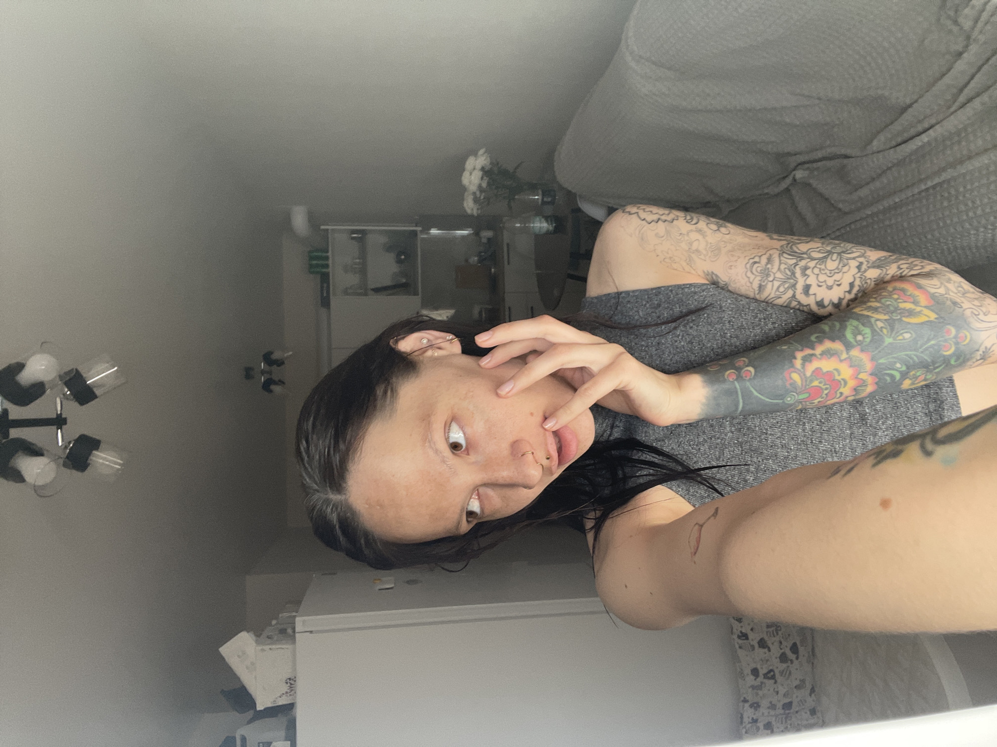 A woman is in an indoor environment. She has dark hair, brown eyes, and fair skin. She has several tattoos on her arms. She is wearing a gray shirt. There is a couch behind her, and a white cabinet or counter below her. There is a light fixture on the wall, and a shelf with flowers is visible in the background. Location: Unknown, Unknown.