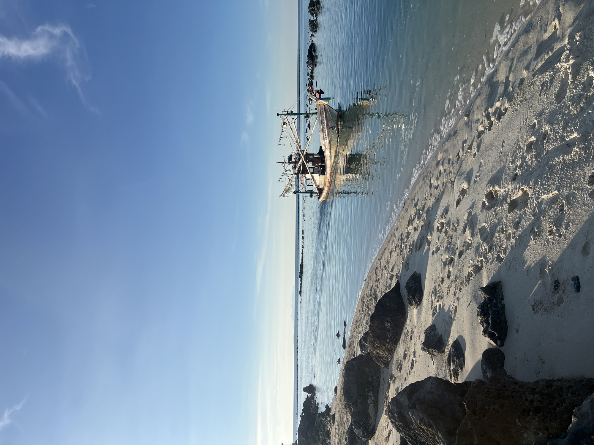 This is a vertical, eye-level, medium shot of a fishing boat on the calm sea near the shore of Koh Phangan, Thailand. The clear blue sky, with a few wispy clouds, takes up the top half of the image. The calm, blue water of the sea occupies the middle section, reflecting the boat and the sky. The foreground is a sandy beach with scattered rocks. A white fishing boat with a multi-colored flag and fishing equipment attached to poles is moving from left to right in the middle ground of the water. A person is visible on the boat. The beach is composed of light-colored sand with footprints and several large, dark rocks scattered along the shoreline. In the far distance, the horizon shows a faint landmass.