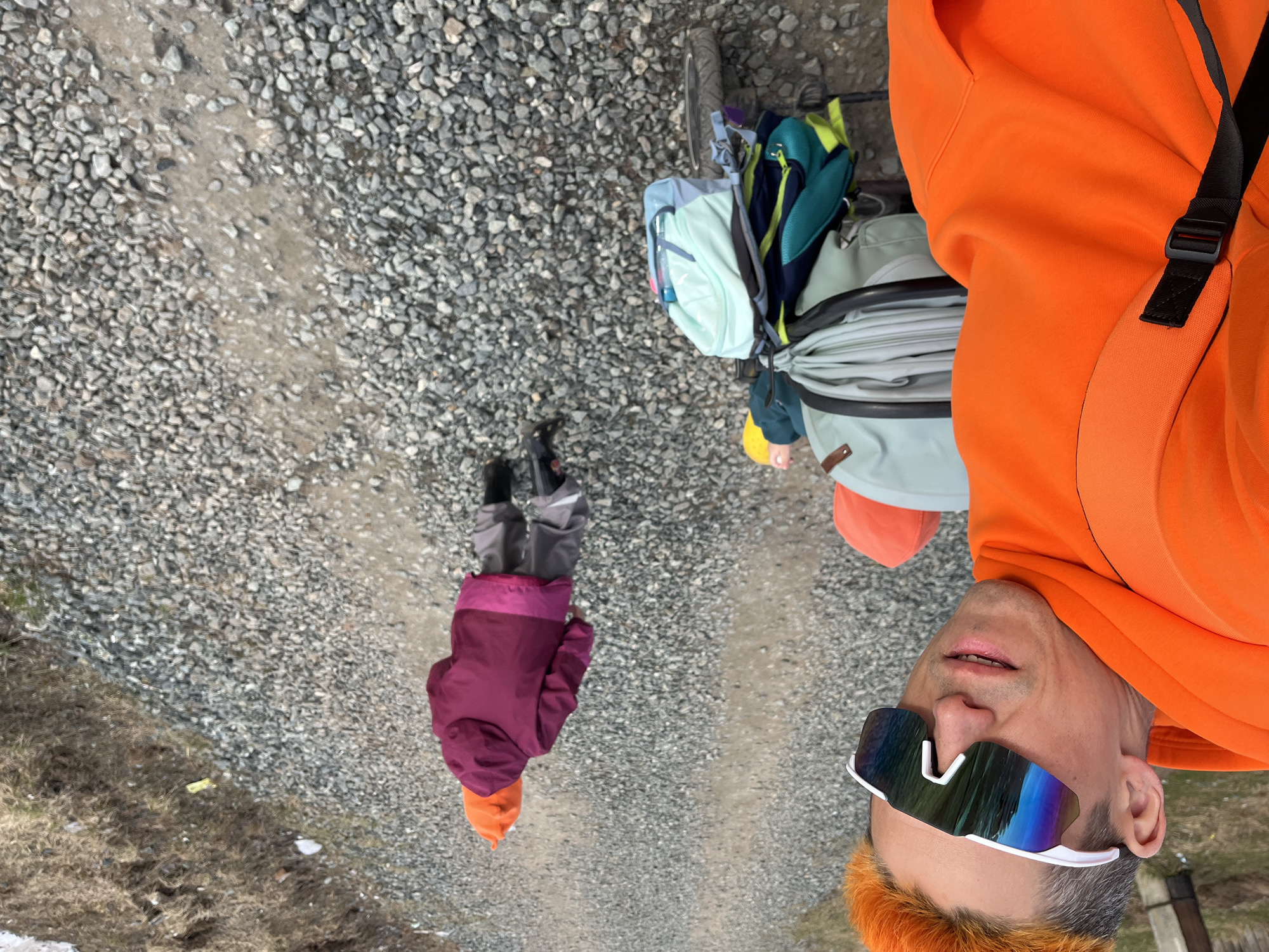 The image captures an outdoor scene in Chaikovsky, Russia, featuring an adult and a child on a gravel path, suggesting an excursion or journey. An adult, visible from the chest up and taking a selfie, wears a bright orange hooded sweatshirt and striking white-framed, multi-colored reflective sunglasses. Their hair appears to have orange highlights. Ahead and further down the path, a child is seen from behind, dressed in a purple jacket, grey pants, and a vibrant orange knit hat.
The setting is a wide, unpaved path covered in light grey gravel. Alongside the path, patches of dry, brown grass and dirt are visible, suggesting an early spring, late autumn, or generally dry conditions. A bicycle, loaded with several backpacks and bags in various shades of light blue, teal, green, and grey, is positioned near the adult, indicating it is part of their travel equipment.
The photograph is taken during the daytime under natural light, likely an overcast sky given the soft illumination. The scene conveys a sense of outdoor activity, such as a bike tour, a hike, or a family outing. No visible text is present in the image.