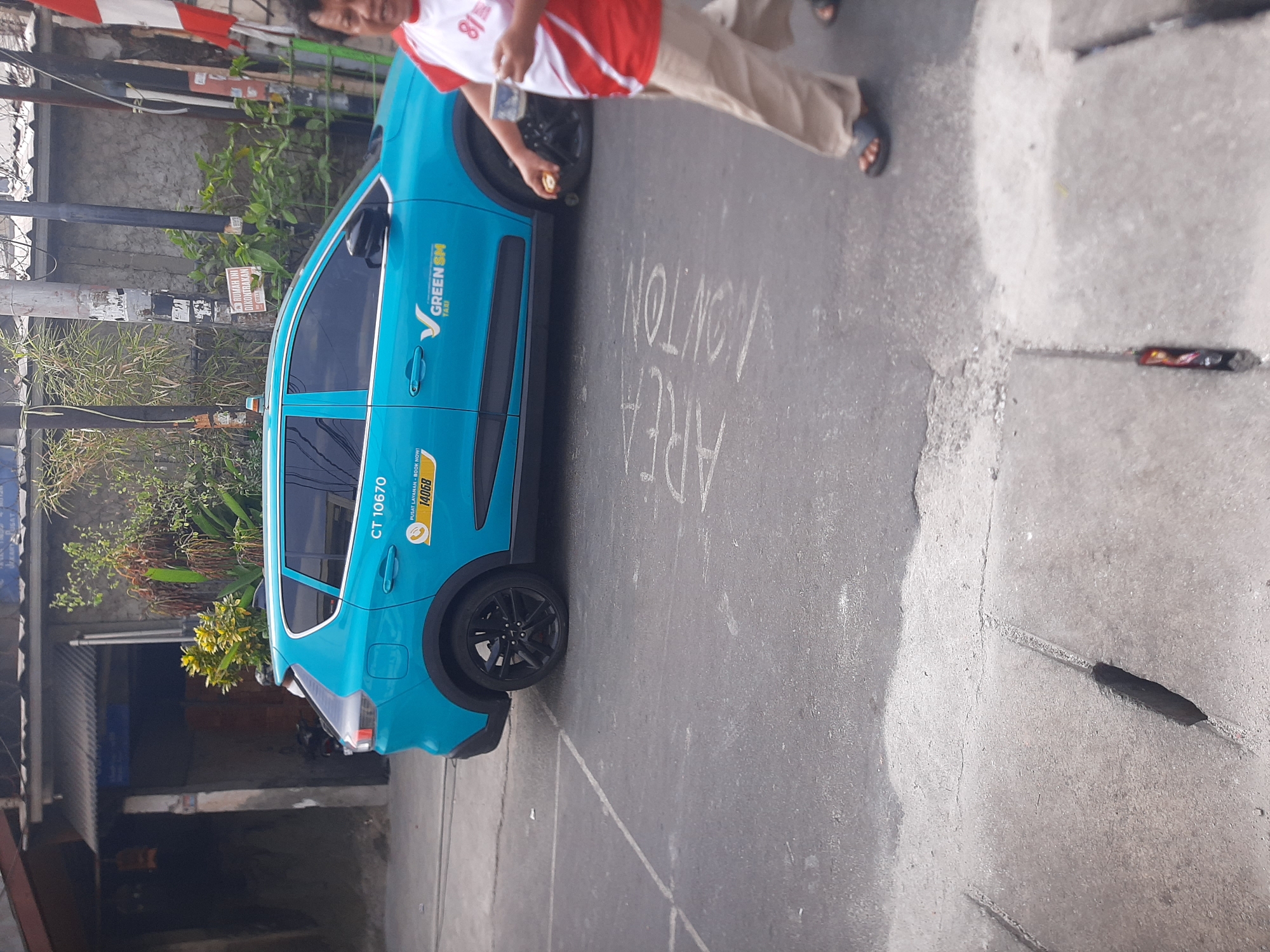 A turquoise taxi is parked on an asphalt road in Central Jakarta, Indonesia. The taxi has the number "CT 10670" on its side and the "GREEN SM TAXI" logo. A person wearing a red and white shirt and beige pants walks past the taxi on the right side of the image, holding a cup in their right hand. The ground is marked with chalk writing that appears to say "AREA LONTON." There are drain covers on the road and buildings in the background.