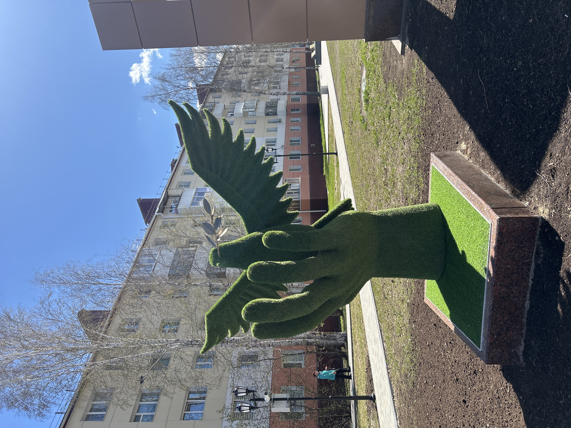 The image shows an outdoor public space in Uray, Russia, featuring a topiary sculpture representing two hands holding a dove with an olive branch. The sculpture is covered in artificial turf and sits on a rectangular base, also lined with artificial turf and edged with dark stone. Behind the sculpture is a multi-story building with light-colored facades and rows of windows. There is a sidewalk and areas of grass and bare soil surrounding the sculpture. A person wearing a blue shirt is standing near a lamppost in the background. The sky is visible, with some trees and clouds. The scene appears to be taken on a sunny day.