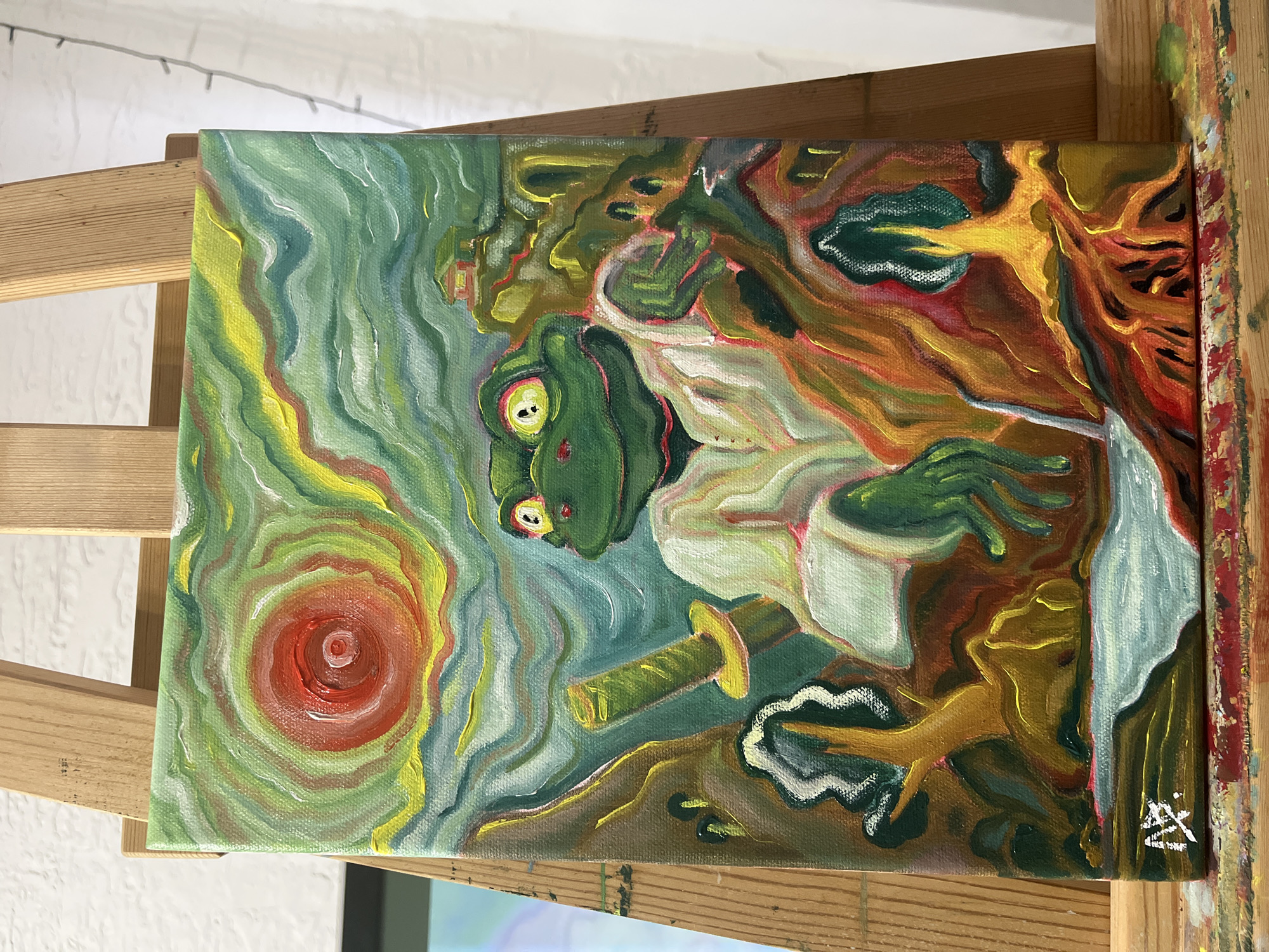 The image shows a painting on a canvas that's placed on a wooden easel. The painting features a green frog with large eyes, dressed in a white kimono-like garment and wielding a sword. The background includes swirling patterns of green, yellow, and red, suggesting a dynamic or otherworldly environment. A red circle with concentric rings is visible in the lower-left corner. A signature is present in the lower-right corner of the painting. The easel itself is made of light-colored wood and shows some signs of paint splatters. The location of the scene is Unknown, Unknown.