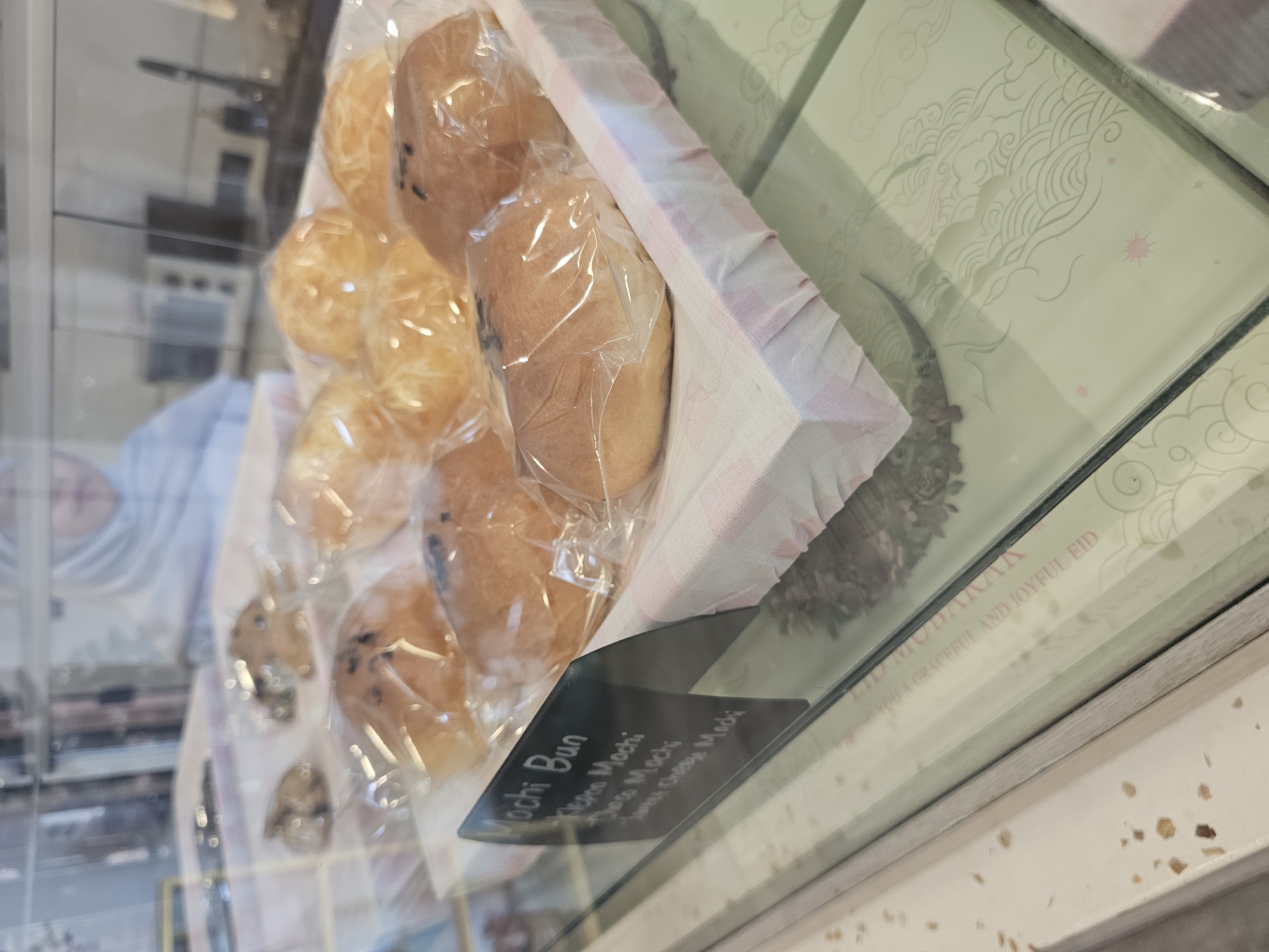 In a display case at a bakery in Banyuurip, Indonesia, an assortment of individually packaged mochi buns are presented. The buns are golden brown and some appear to have dark speckles, possibly chocolate chips or sesame seeds. A black sign in the foreground reads "Mochi Bun," followed by "Aka Mochi," "Choco Mochi," and "Sweet Cheese Mochi." The buns are arranged on a white and pink checkered cloth. In the background, a person wearing a light-colored hijab is partially visible, and behind them, a blurred view of a kitchen or serving area can be seen. The display case reflects the surrounding environment, including what appears to be patterned boxes with the text "HIKMAT MODAIKAN" and "GRACEFUL AND JOYFUL EID." The overall scene depicts a retail setting focused on baked goods.