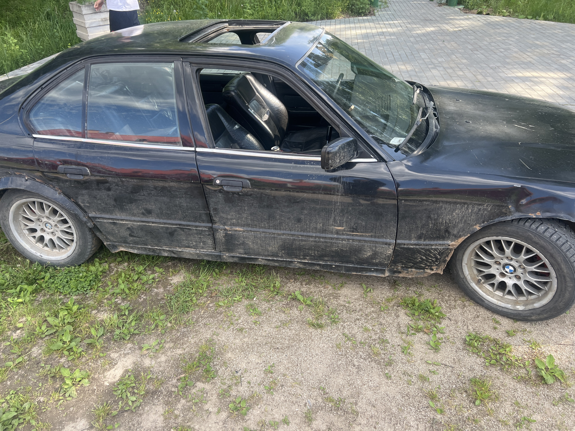The scene depicts a black BMW sedan, likely an E34 model, parked outdoors in Noviy, Russia, on a dirt and gravel surface with sparse patches of green grass and weeds. The vehicle exhibits significant external wear and grime, with thick layers of mud and dirt adhering to the lower body panels, doors, and fenders, particularly evident on the driver's side. Visible rust and body damage are present on the rear driver's side fender arch. The front hood displays multiple scratches and indentations, and the windshield wipers appear aged. Both the driver's side rear wheel and the front passenger side wheel are multi-spoke alloy rims, heavily soiled, with the BMW logo visible on the front wheel. The front passenger side tire appears to be a winter tire. The car's sunroof is open. Through the windows, the interior shows worn black leather seats. A can is visible on the floor or passenger seat. In the background, to the right of the car, a paved area with interlocking grey pavers is visible. Further back and to the left, green foliage and a person in a white t-shirt and dark pants standing near a light-colored rectangular object are partially discernible. The lighting indicates daytime conditions.