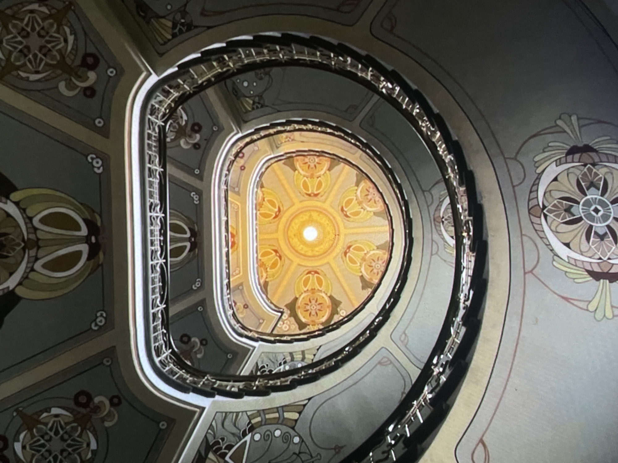 This is a low-angle, eye-level shot looking up into a spiral staircase of a building in Harmanli, Bulgaria. The staircase is ornate, with decorative elements on the walls and ceiling. The ceiling is illuminated from above, creating a bright focal point at the top of the stairwell. The walls feature geometric patterns and stylized floral motifs in muted colors like grey, beige, and yellow. The railings of the staircase are dark and appear to be made of metal. No people are visible in the image. The environment suggests an older, well-maintained building with an artistic architectural design.