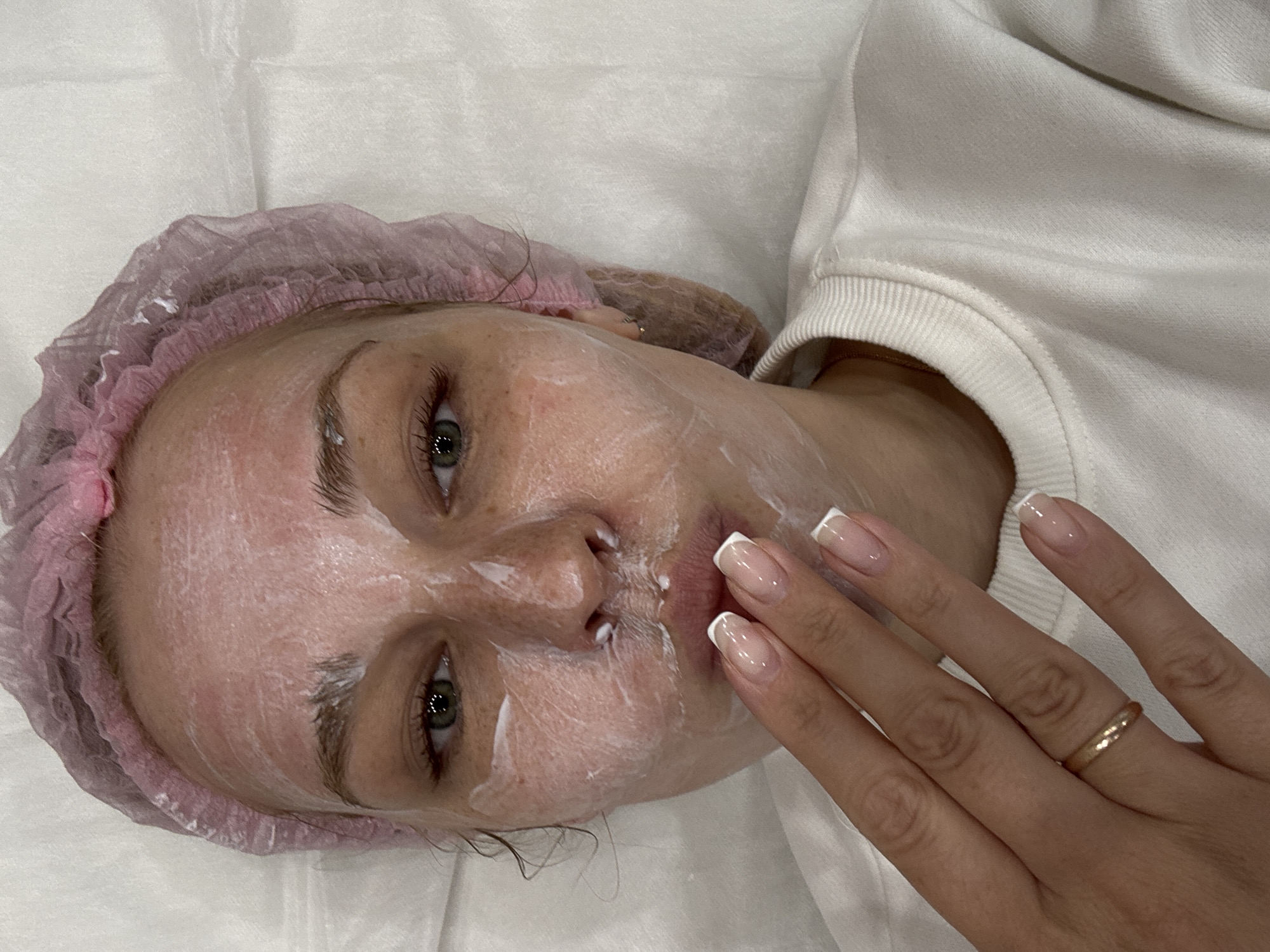 A woman is lying on her back, wearing a pink hair net and a white sweater. Her face is covered in a white cream, likely a facial mask. Her right hand is near her face, with neatly manicured nails. She is wearing a gold ring on her ring finger. She is lying on a white surface. The location is possibly in Rusanovo, Russia.