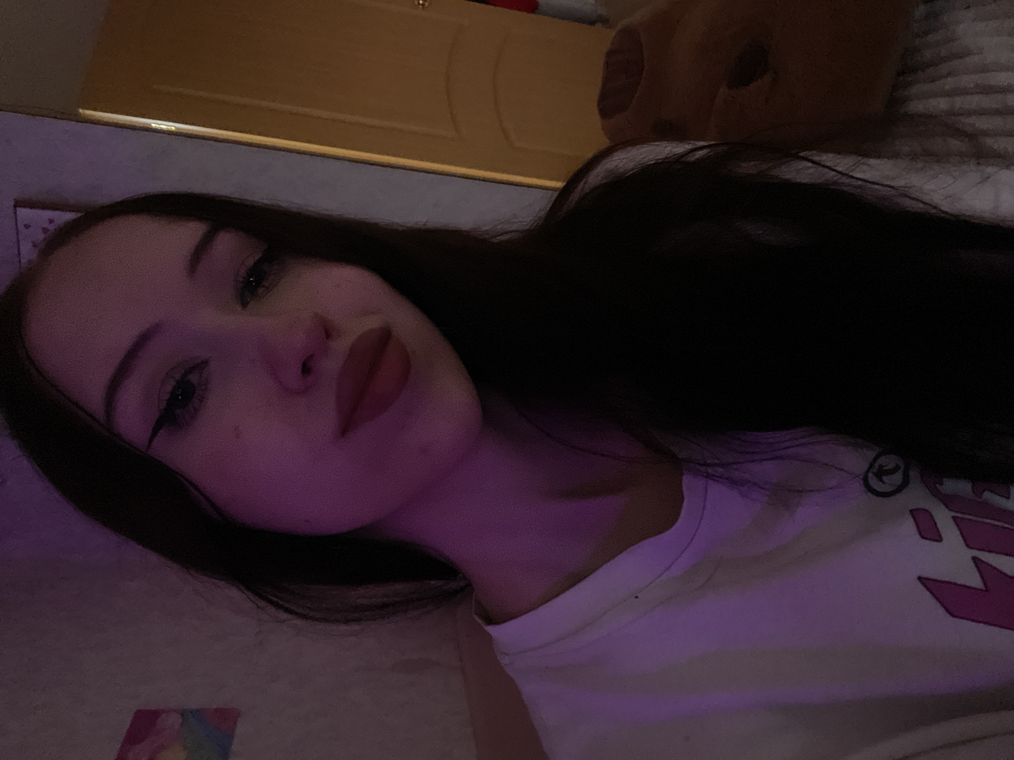 This is a selfie taken by a young woman in what appears to be a bedroom. The lighting is dim and colored with purple hues, likely from ambient lighting or a mood lamp. The woman has dark hair, winged eyeliner, and is wearing a white t-shirt with pink lettering. To her left, a small colorful painting is visible on the wall, and to her right, a doorway or closet door is partially open, revealing a warm, golden light. The overall impression is of a casual, personal moment captured in a domestic setting. There is no information in the image to suggest it is located in Votkinsk, Russia.