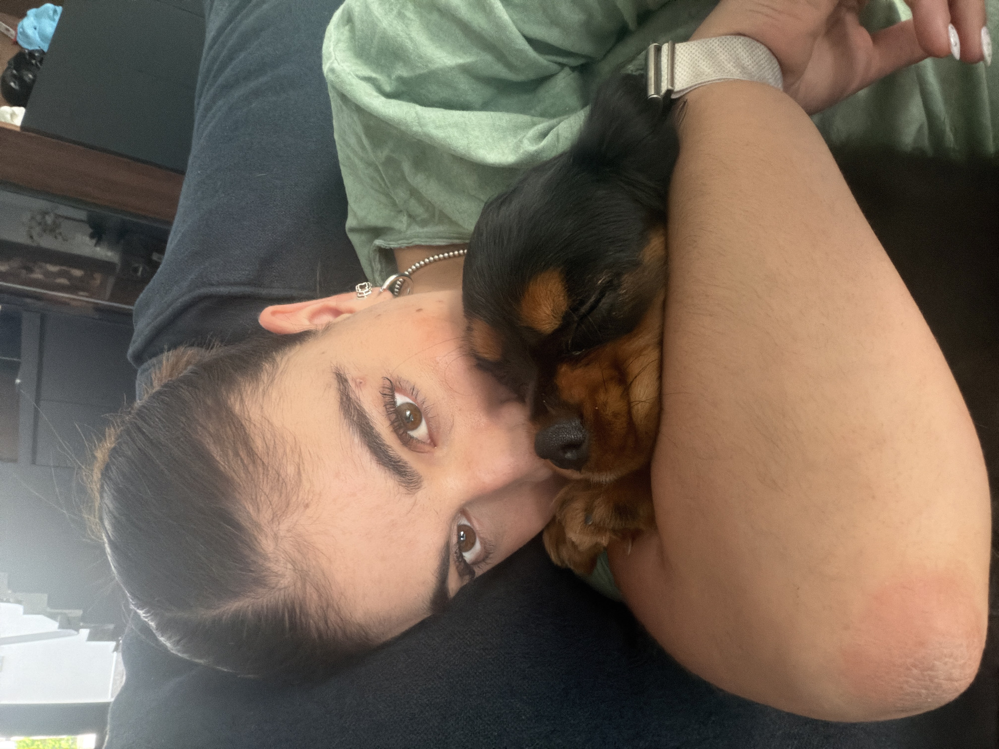 A woman is lying down and holding a small black and tan dog, possibly a Cavalier King Charles Spaniel. She is looking directly at the camera. She is wearing a light green shirt and has a silver watch on her wrist. Her hair is pulled back. The dog is nestled against her arm, and its eyes are closed. The location is not determinable from the image.