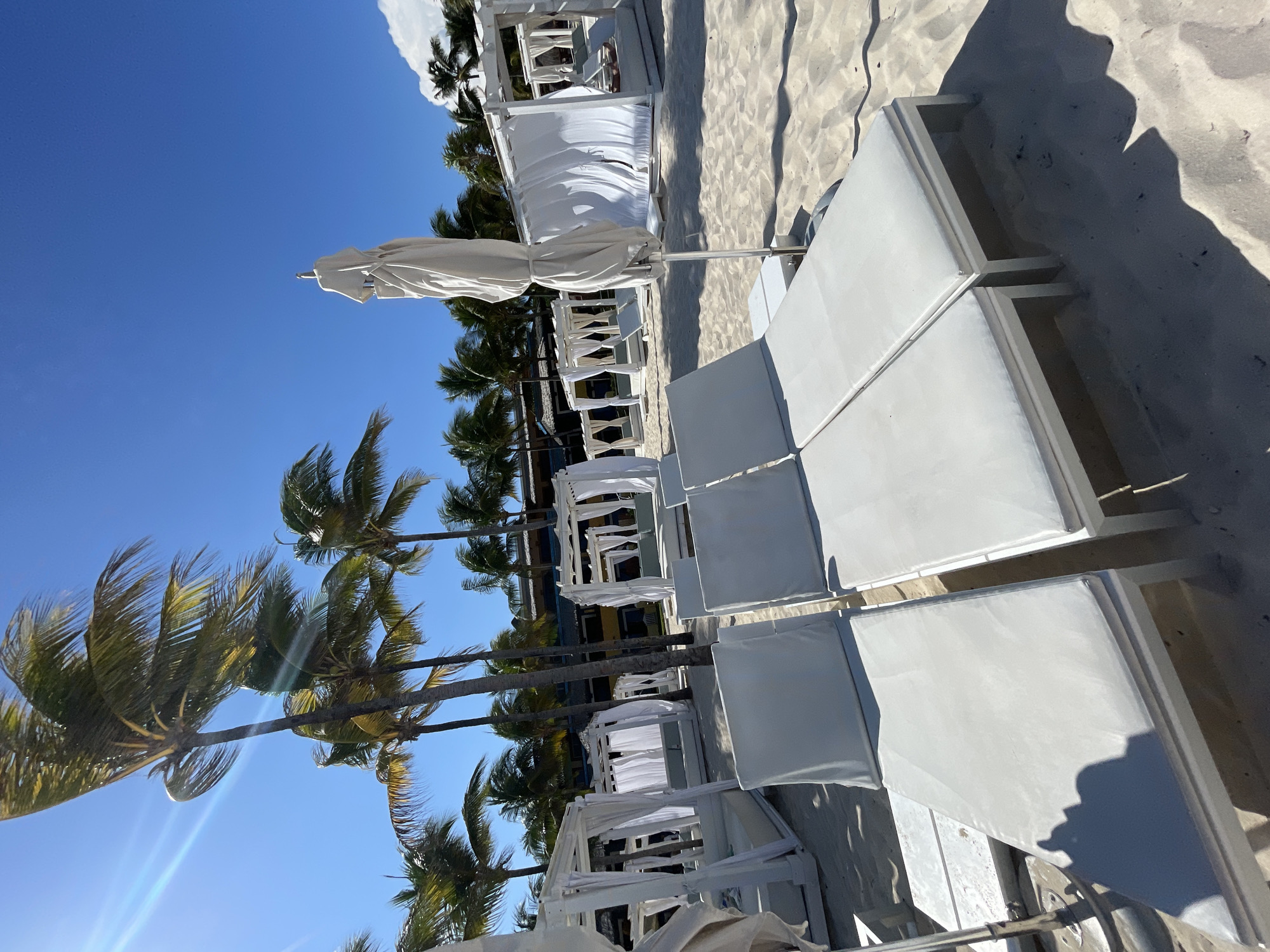 The image shows a beach scene in Playa del Carmen, Mexico. Rows of white lounge chairs and cabanas line the sandy shore under a clear blue sky. Palm trees are visible along the beach. There are no people visible in the image.