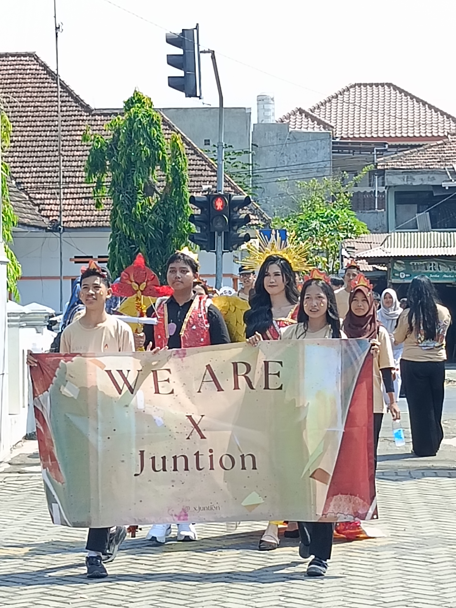The image depicts a parade or procession in the city of Kediri, Indonesia. In the foreground, a group of people are holding a banner that reads "WE ARE X Juntion". The banner is colorful and appears to be made of fabric. Several individuals are visible behind the banner, some wearing decorative headwear, suggesting a festive occasion. A woman with a golden halo-like headdress stands prominently. In the background, there are buildings with tiled roofs, trees, and traffic lights, indicative of an urban setting. One traffic light displays a red signal for pedestrians. The ground is paved with interlocking bricks. The overall scene suggests a community event taking place outdoors on a sunny day.