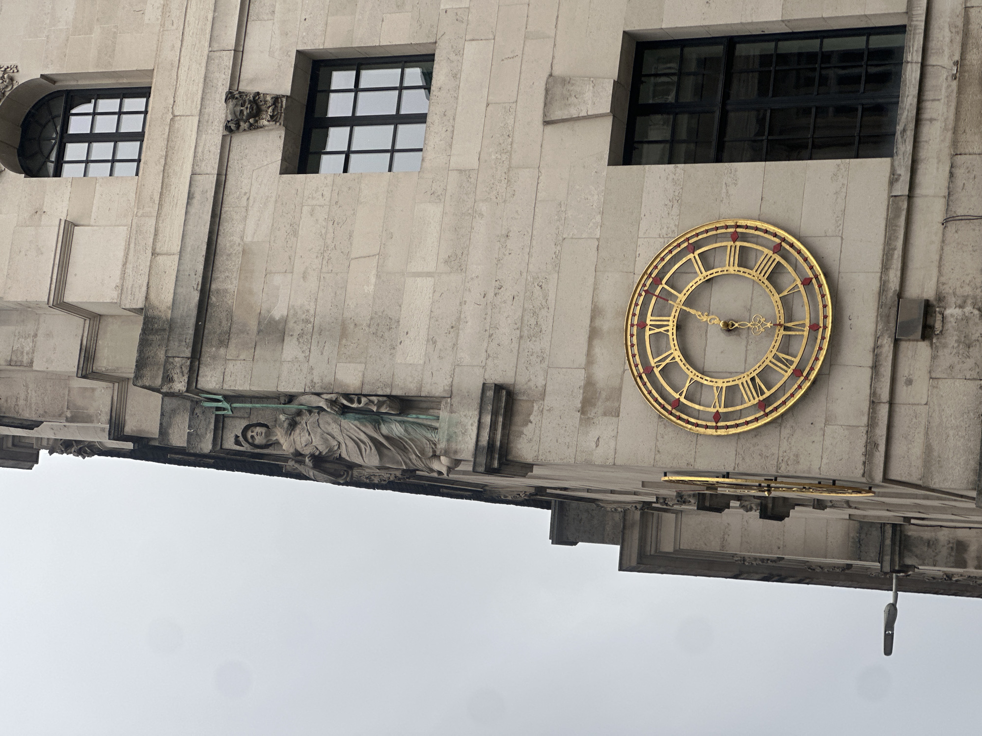 The image depicts a section of the Senate House in London, United Kingdom. It shows a portion of the building's exterior featuring several windows, a prominent clock face with gold numerals and hands, and a sculpture of a woman with a spear. The building's facade is made of light-colored stone. The clock indicates that it is between 10:00 and 10:30. The sky is overcast and gray.