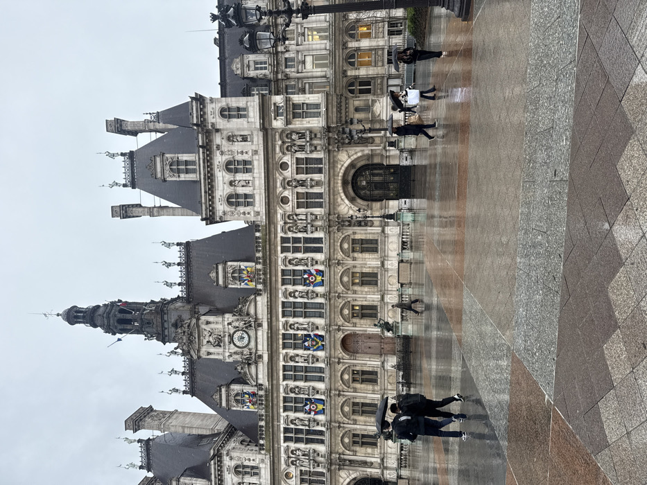 A tall, ornate building occupies the left side of the frame. It features intricate architectural details, including multiple windows, statues, and a clock. Several flags adorn the building's facade. On the right, a large square paved with geometric patterns of different colors extends toward the building. People are walking, and their reflections are visible on the wet pavement, suggesting it has recently rained.