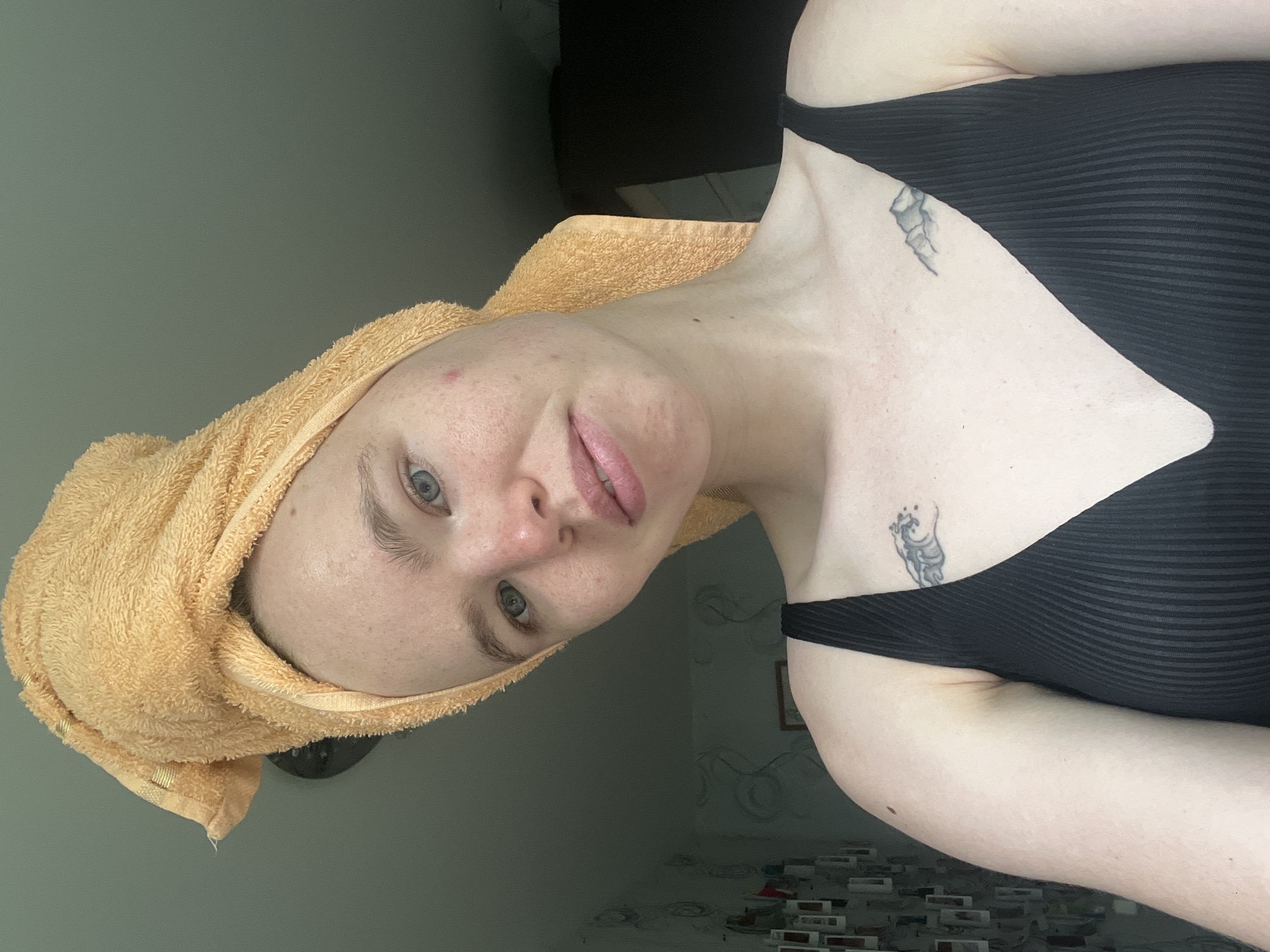 A person is depicted in a medium close-up, indoors, with their head wrapped in a light orange, terry cloth towel, suggesting they may have recently showered. The individual has light skin, light blue-grey eyes, and light eyebrows. They are looking directly at the camera with a neutral expression, possibly a slight smirk, and their lips are lightly colored. Visible skin details include freckles and a few red blemishes on the forehead and cheeks.
The person is wearing a black, ribbed tank top or camisole. Two small, faded tattoos are visible on their left shoulder/upper chest area: one appears to be a feathery or leafy design near the collarbone, and another, more abstract or stylized creature-like design, is situated lower on the chest, near the armpit.
The background consists primarily of a plain, muted greenish-gray wall. To the right, behind the person's shoulder, a darker, blurred area is visible, possibly part of furniture or a doorway. Further back and to the right, blurry decorative elements, including what might be a framed picture and some patterned surfaces (possibly tiles or wallpaper), can be seen. The image is taken indoors in Chaikovsky, Russia. The lighting is soft and even, typical of indoor settings. No visible text is present in the image.