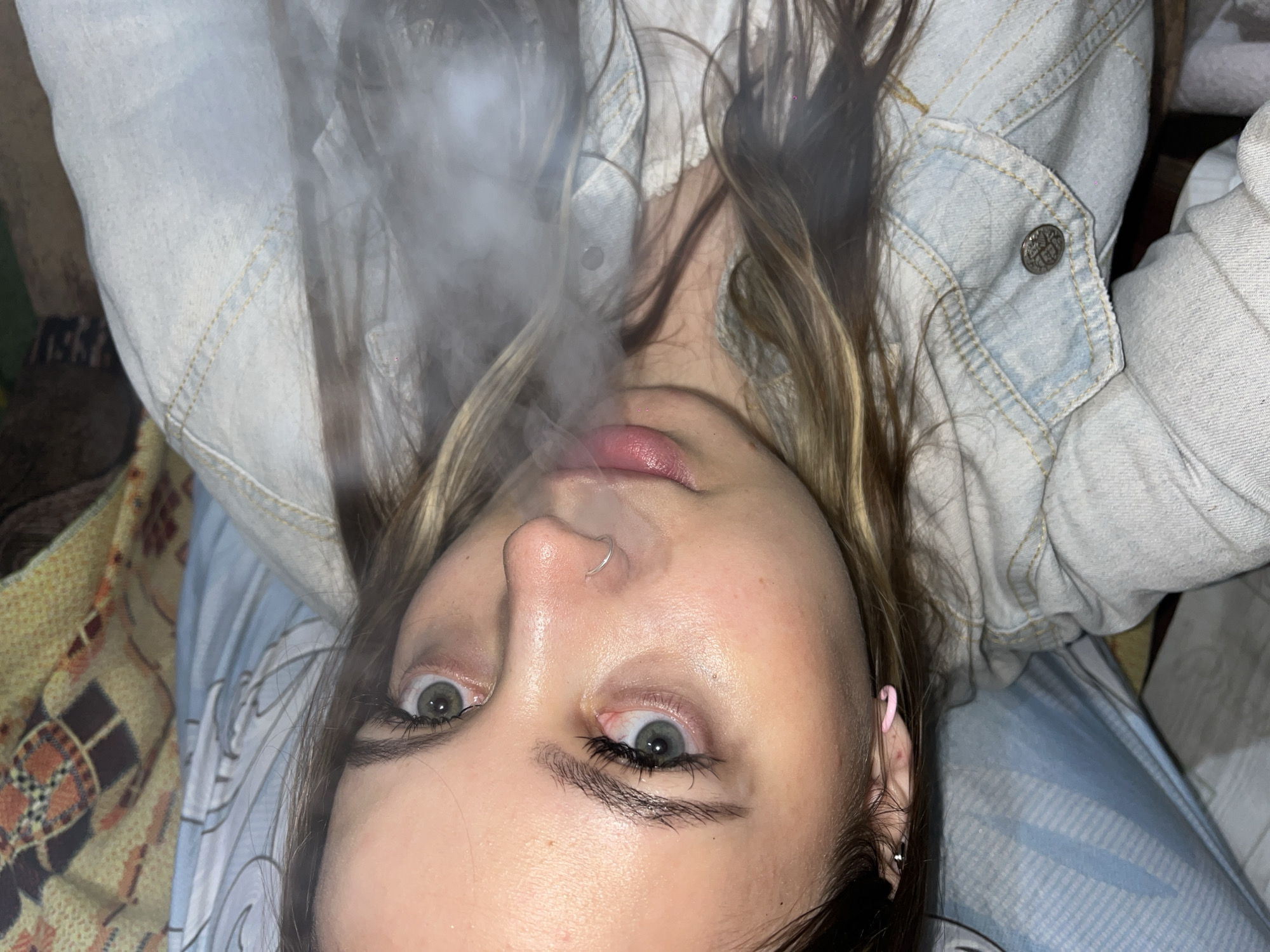A young woman with light skin, light brown hair with blonde streaks, and green eyes is lying on her back, exhaling smoke. She is wearing a light blue denim jacket and a white lace-trimmed top. She has a nose ring and is wearing earrings. The background includes a blue plaid blanket and a patterned object with shades of brown. There is no indication that this is in Novopokrovskaya, Russia.