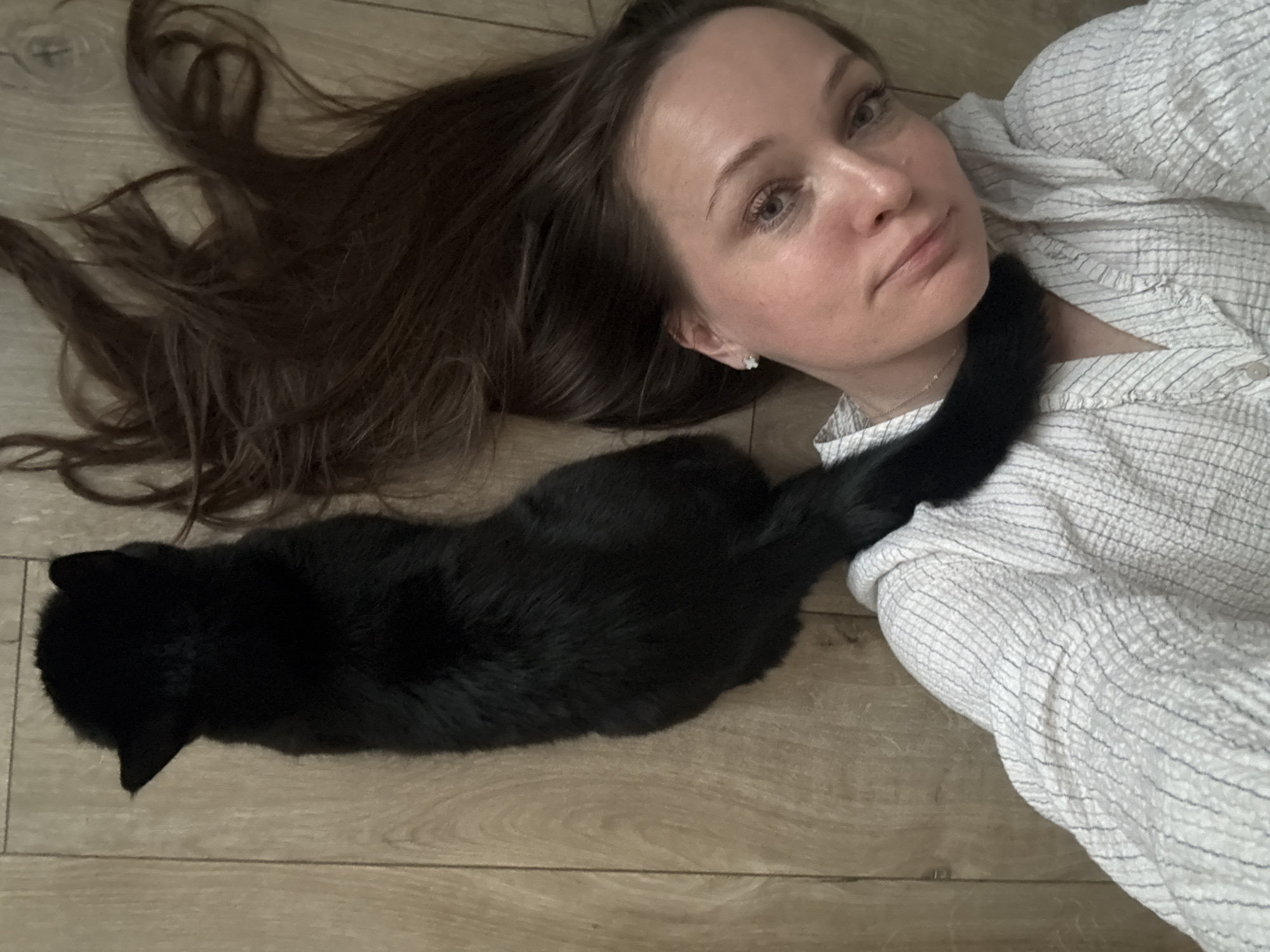 A woman and a black cat are lying on a light-colored wooden floor. The woman has long brown hair spread out on the floor next to her head. She is wearing a white shirt with light grey stripes. The black cat is lying next to the woman, its tail resting on her shirt. The location is likely a residence, based on the interior elements. The city of Chaikovsky, Russia, cannot be confirmed from this image.