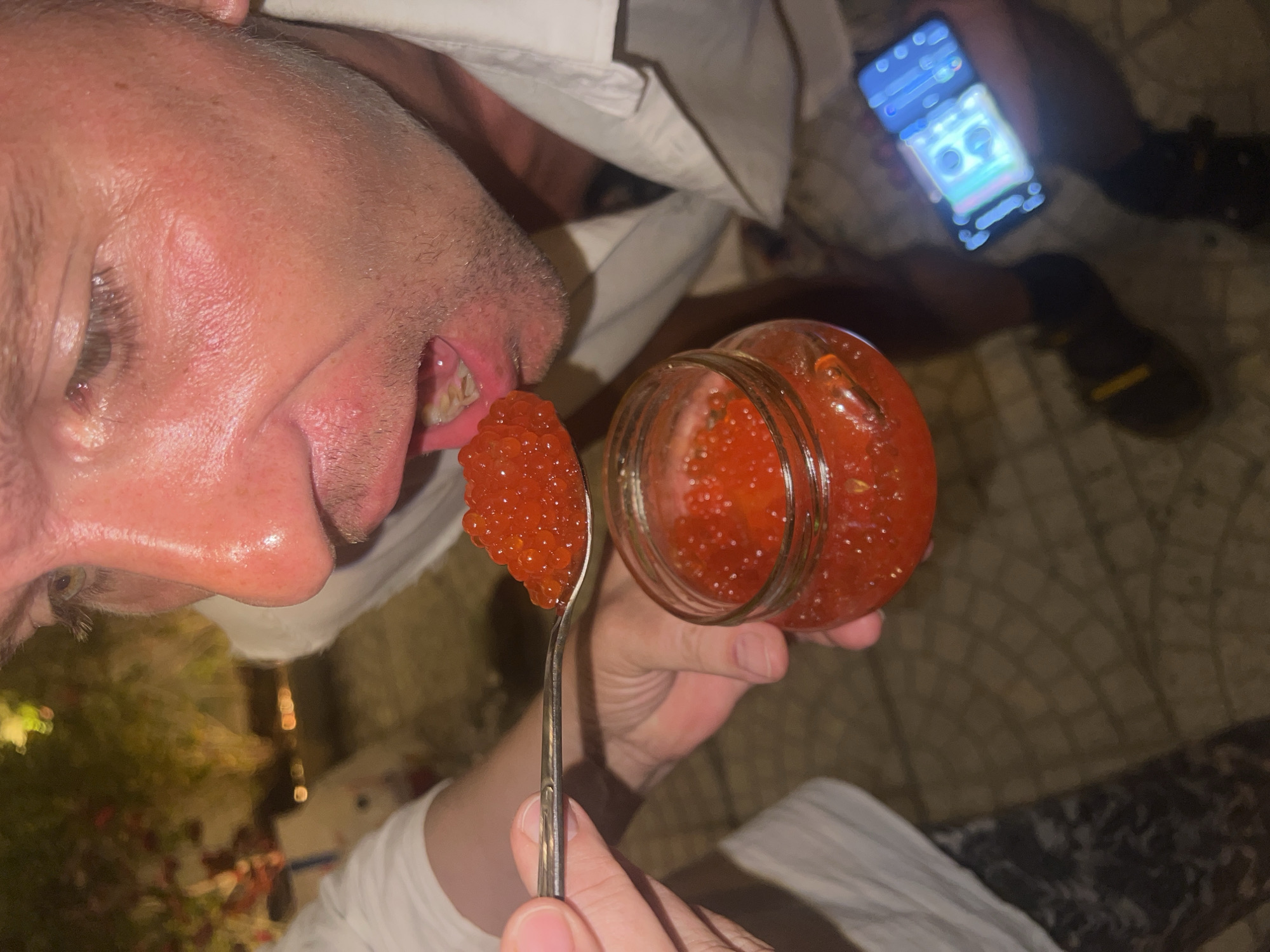 A close-up shot captures a person about to eat a spoonful of red caviar from a glass jar. The individual is leaning forward, with their mouth open, as a spoon laden with caviar is brought towards it. The person is wearing a white collared shirt. In the background, out of focus, there's greenery and some lights. To the right, a person's hand holds a smartphone displaying a bright blue interface. The ground appears to be paved with square tiles. The scene seems to be outdoors, possibly at night. There is no indication of the location being in Đà Nẵng, Vietnam.