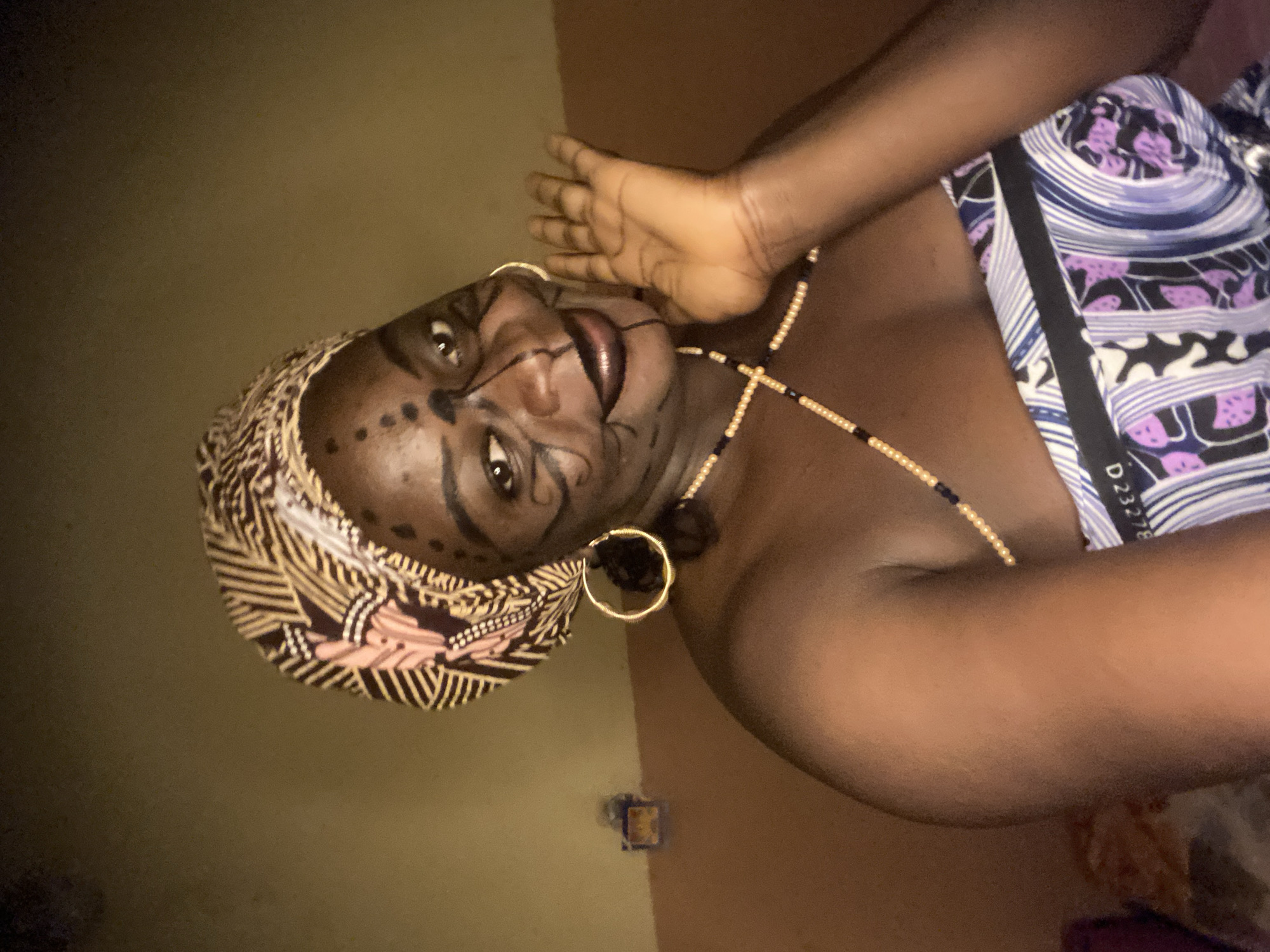 A medium shot depicts a person with dark skin, presumably female, with decorative facial markings and wearing a patterned headscarf and beaded necklace, suggesting traditional or cultural attire. The person is positioned on a wall, facing the camera with a slight tilt. The background appears to be a dimly lit interior, with a plain wall and minimal visible details. The location appears to be in Maigizo, Nigeria, based on prior context. The person's expression is neutral, and there are gold hoop earrings visible. The clothing includes a patterned garment with shades of purple, white, and black. The lighting is subdued, casting shadows and impacting overall visibility.