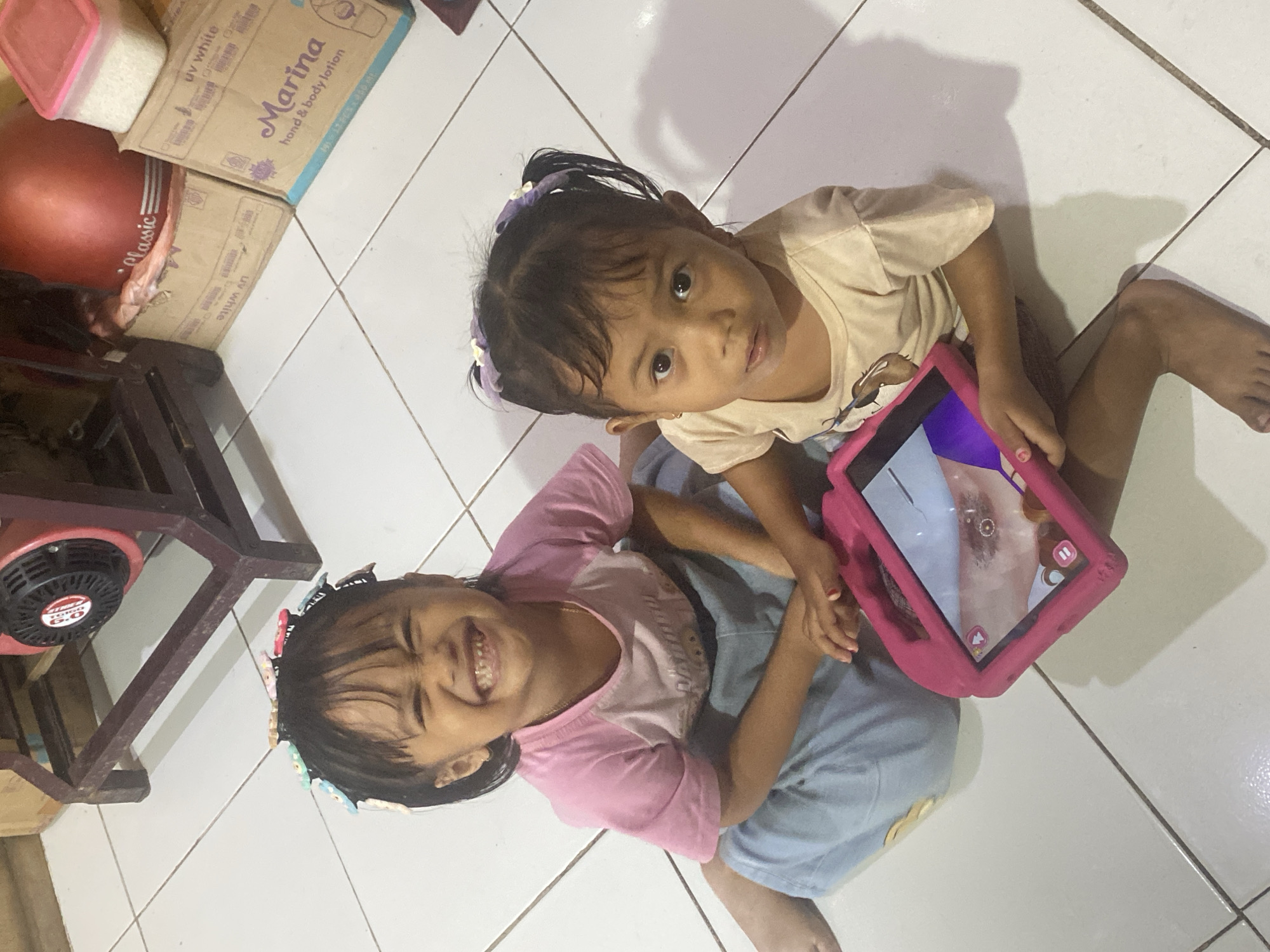 The image depicts two young girls sitting on a tiled floor in what appears to be a residential setting. The girl on the left, wearing a pink shirt and blue pants, is smiling broadly, her eyes closed. Her hair is adorned with colorful hair clips. The girl on the right, in a light-colored t-shirt, is looking up with a neutral expression. She is holding a pink tablet, which is displaying an image of what looks like a person's face, possibly a close-up of an eye. In the background, there is a red and black machine, cardboard boxes, and a container of white rice. The overall environment suggests an indoor space, likely a home. Based on the visible text on the boxes, which includes "Marina hand & body lotion" and "uv white," it's possible the location is a household in Indonesia, potentially in Ta', a city known for its commercial and industrial activities.