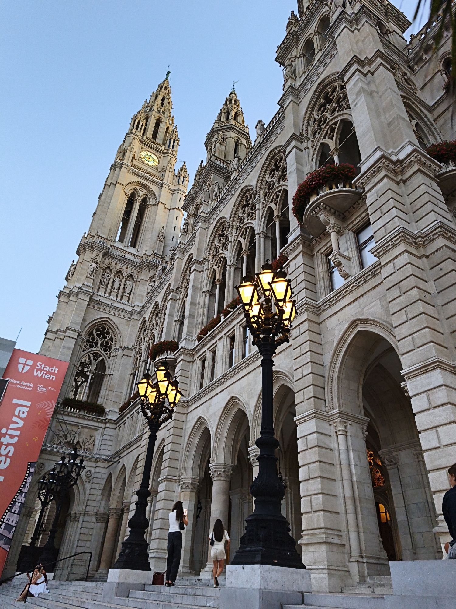 The image shows the Rathaus (City Hall) in Vienna, Austria, viewed from a low angle. The grand Gothic Revival building features intricate stonework, ornate windows, and towering spires. In the foreground, a tall, black lamppost with glowing yellow lights stands prominently on a wide stone staircase. Several people are present on the stairs. Two women are walking up the stairs, one of whom is holding a phone. Another woman is sitting on the steps. A red banner with the text "Stadt Wien" (City Vienna) and other inscriptions is visible on the left side of the image. The sky is clear and blue, indicating daylight.
