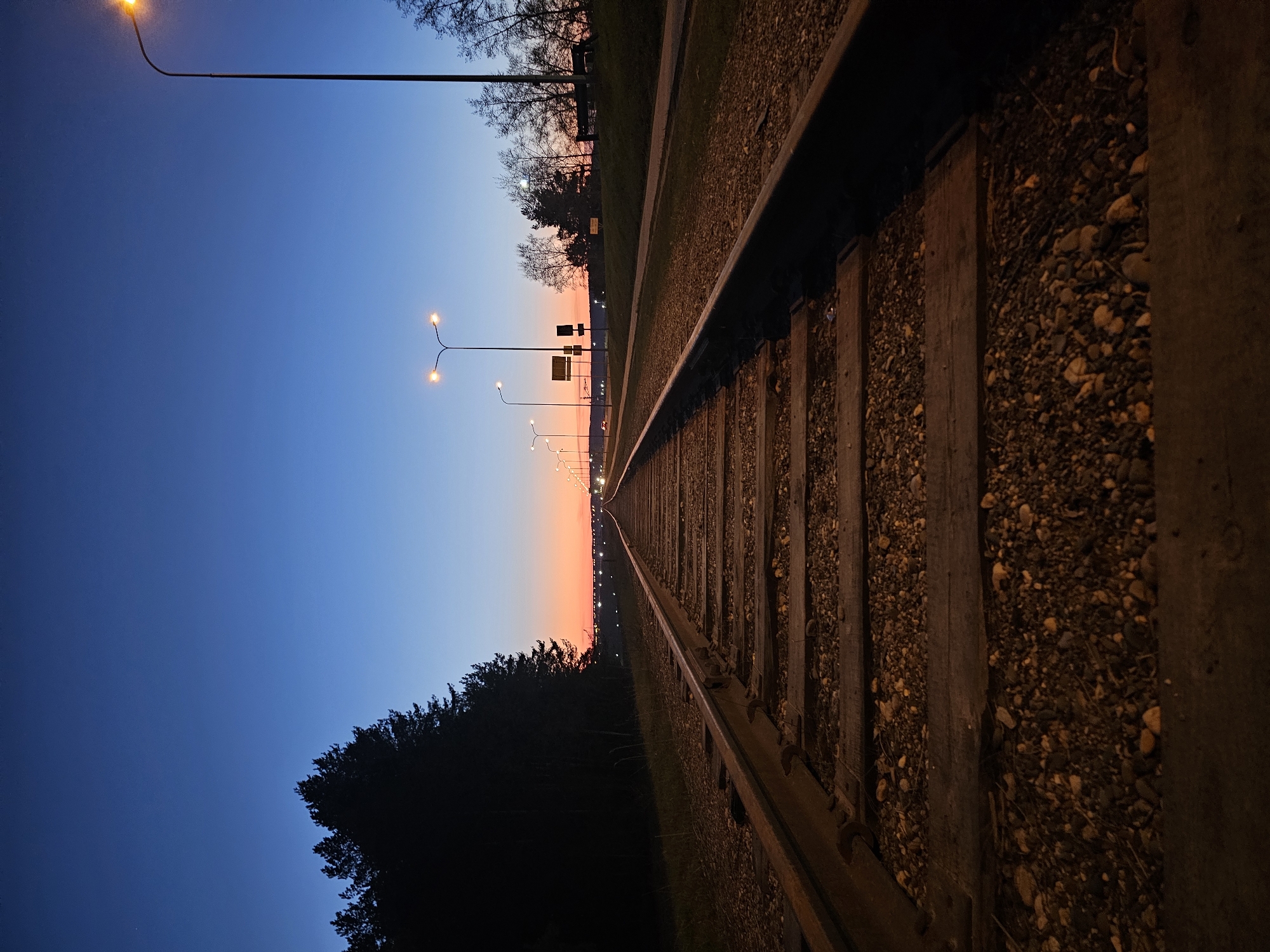 The image shows a section of railroad tracks at dusk in what appears to be the city of Chaikovsky, Russia. The railroad tracks recede into the distance, flanked by gravel and wooden ties. Streetlights line the side of the tracks. The sky transitions from a deep blue on one side to orange on the horizon. Trees and vegetation are visible along the edges.