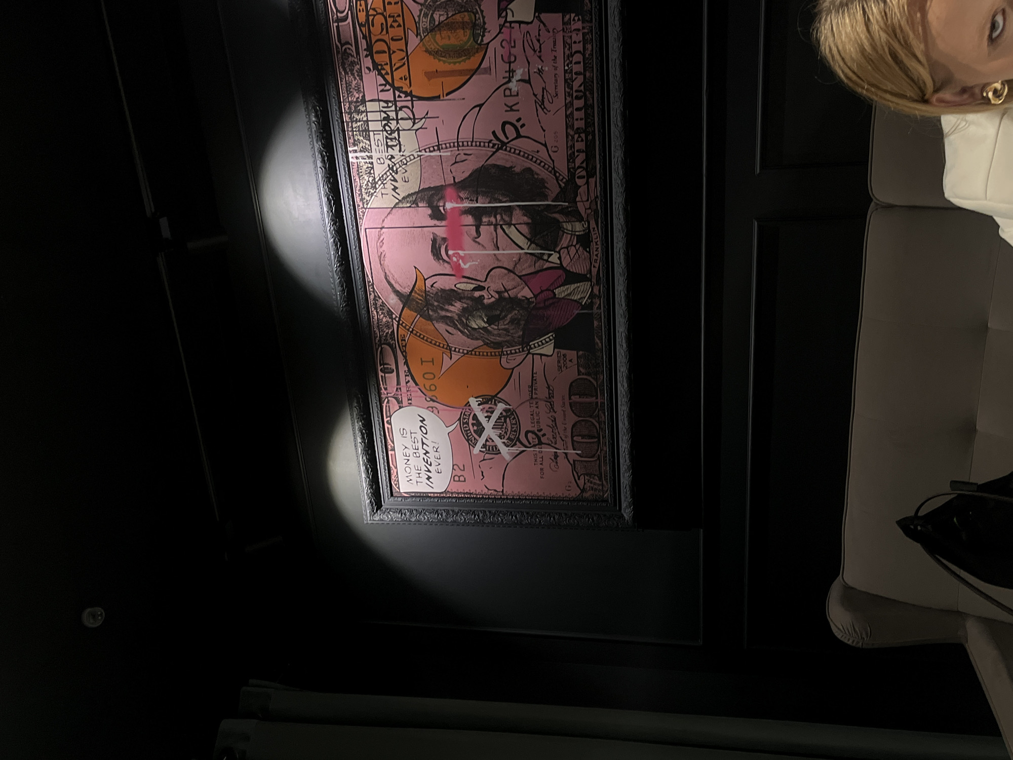 The image captures an indoor scene, likely a dimly lit room in Chaikovsky, Russia, dominated by a striking framed artwork. The art piece is a pop-art style depiction of a one-hundred-dollar bill, primarily in vibrant pink and orange with black outlines and graffiti-like additions. The central figure, a stylized Benjamin Franklin, has a bold red stripe across his eyes and cartoonish devil horns. A speech bubble within the artwork reads: "MONEY IS THE BEST INVENTION EVER!". Other visible text includes "ONE HUNDRED", "FRANKLIN", "THIS NOTE IS LEGAL TENDER FOR ALL DEBTS PUBLIC AND PRIVATE", "Secretary of the Treasury" (partially obscured), "SERIES 2006 A", "B2", "G 205", and "3.KP46279".
A focused spotlight highlights the upper left section of the artwork. To the right, a person with blonde hair is partially visible, wearing a white top and a prominent gold hoop earring. They appear to be leaning or sitting on light-colored upholstered furniture, which can be seen in the bottom right. The background consists of dark walls or paneled doors, creating a somber yet sophisticated atmosphere. No specific activity is taking place beyond the implied observation of the artwork. The scene suggests a modern interior, possibly a lounge or gallery, with a focus on contemporary art.