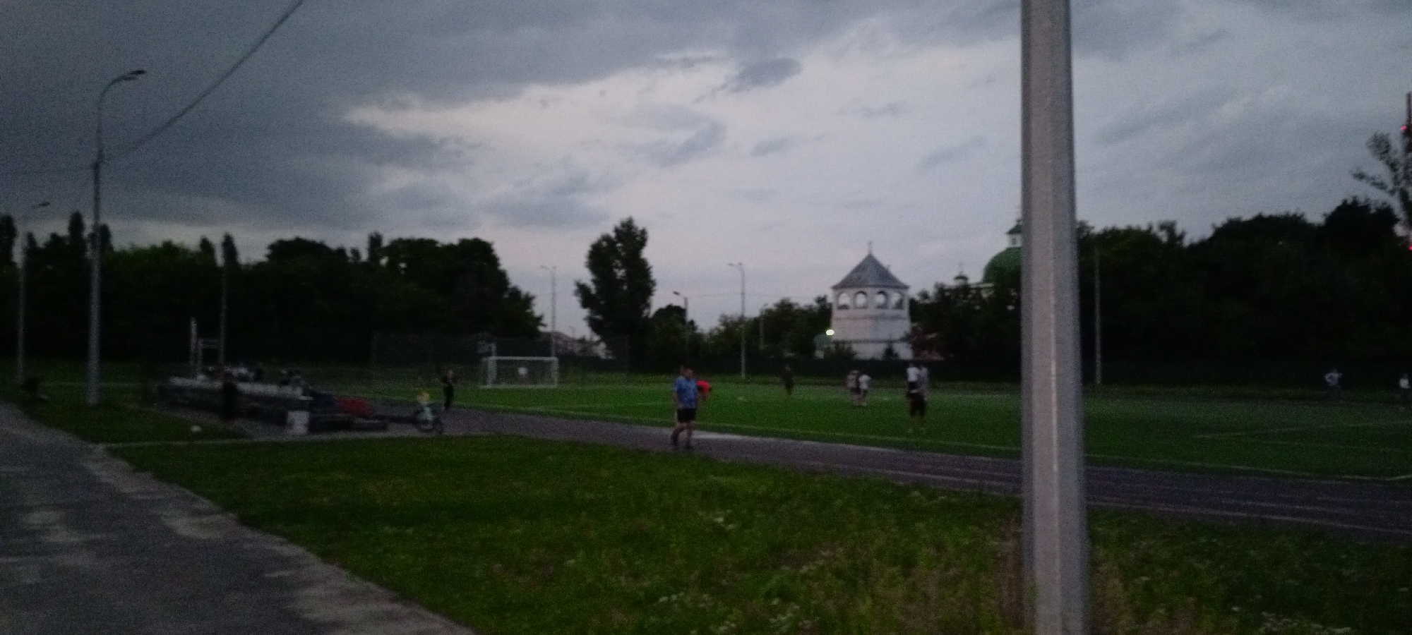 The image captures an outdoor scene in Kirsanov, Russia, during what appears to be twilight or overcast conditions. In the foreground, a gray, textured pathway runs diagonally across the lower left of the frame. Adjacent to this is a well-maintained green grassy area. Further back, a running track is visible, and beyond that, a sports field, likely a soccer field, is set up with a goalpost on the left side. Several individuals are scattered across the field, engaged in various activities. One person in a blue shirt and dark shorts is walking on the track, holding a red spherical object. Another figure in dark clothing is standing near the goalpost. Several other people are spread out on the grass. In the background, a dense line of dark green trees forms a backdrop, and to the right of the trees, a distinctive white tower with arched openings and a conical roof is visible, suggesting a historical or religious structure within the city. Further to the right, a green dome of a building can be seen peeking above the trees. The sky is overcast with heavy, dark clouds, contributing to the muted light of the scene. Streetlights are also visible, with one prominently positioned on the left side of the image. The overall impression is of a public park or sports complex in Kirsanov.