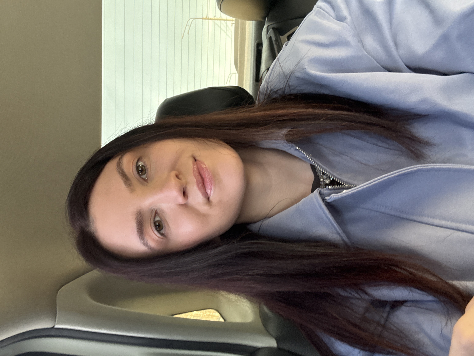 A woman with long, dark hair and fair skin is inside a car. She is wearing a light blue hooded jacket with a zipper. The interior of the car is visible, including the black seat and the car's door panel. Outside the car is a window with vertical blinds, suggesting a building facade. The photograph was likely taken in Kazan, Russia.