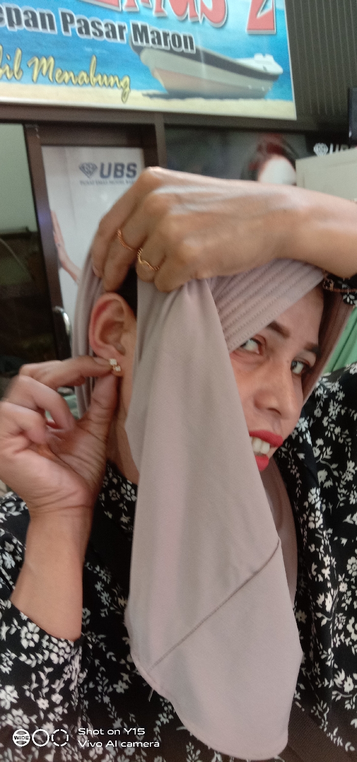 A close-up image depicts a person, likely female, adjusting a small, light-colored stud earring in their left ear with their left hand. Their right hand, adorned with two gold-colored rings, holds back a light purple ribbed headscarf (hijab) that partially covers their face. The visible portion of the face shows red lipstick, exposed teeth, and the left eye looking upwards. The individual wears a black garment with a white floral pattern. In the background, a blue banner is visible with partial text reading "epan Pasar Maron" and "bil Menabung," along with a graphic of a small boat. Below the banner, a framed sign displays the "UBS" logo (a diamond with "UBS") and the partial text "Pusat Emas Murni." The scene is set in a retail environment, likely a gold or jewelry store, located in or near Pasar Maron (Maron Market) in Maron, Indonesia.