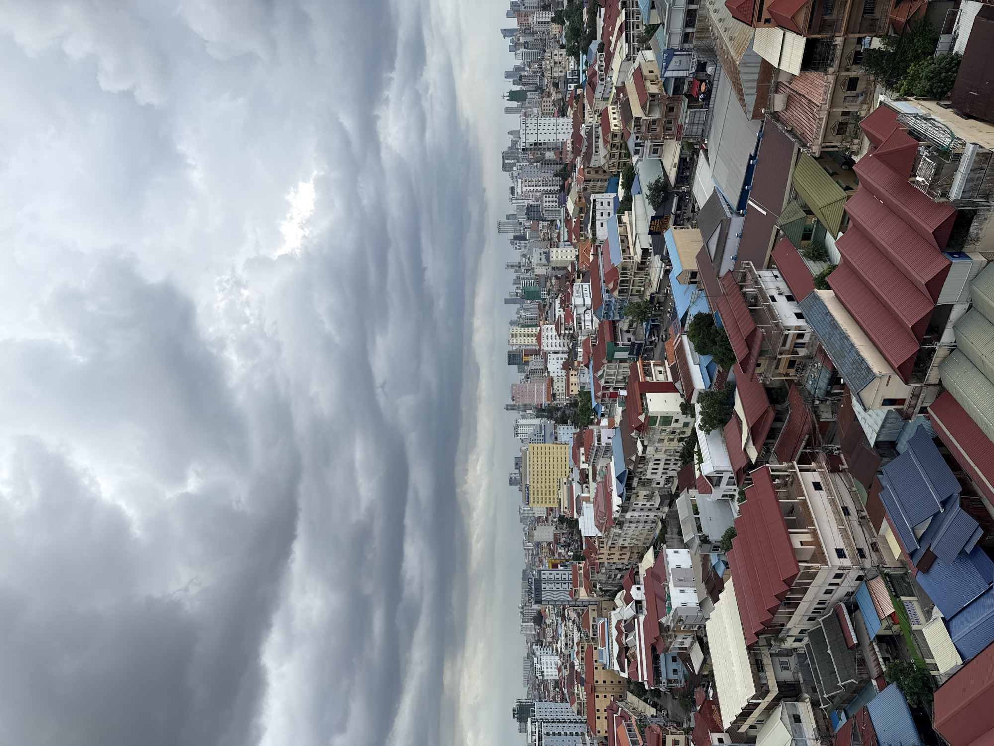This is a high-angle, full-frame, eye-level photograph taken from a building in Khan Mean Chey, Cambodia. The image depicts a dense urban landscape characterized by a variety of buildings, ranging from low-rise structures with red-tiled roofs to taller, modern high-rises in the background. The sky is heavily overcast with dark, textured clouds, suggesting an impending storm or a heavily cloudy day.
In the foreground and midground, numerous residential and commercial buildings are visible. Many of the lower buildings feature vibrant red roofs, often with multiple pitched sections. These structures are tightly packed, with narrow streets or alleys visible between them. Some of these buildings have balconies and multi-story designs.
As the view recedes into the background, the buildings become taller and more modern. A cluster of high-rise buildings, including office towers and apartment complexes, dominates the horizon. These structures vary in architectural style and color, with some featuring glass facades and distinct geometric shapes. The overall impression is one of a rapidly developing urban center.
There are no visible people or animals in the photograph. The scene is static, focusing solely on the built environment and the atmospheric conditions. The overall color palette is dominated by the muted grays of the sky, the reds of the roofs, and the various neutral tones of the building exteriors. The lighting is diffused due to the cloud cover, resulting in soft shadows and a somewhat somber mood. The image captures a typical day in a bustling Asian city.