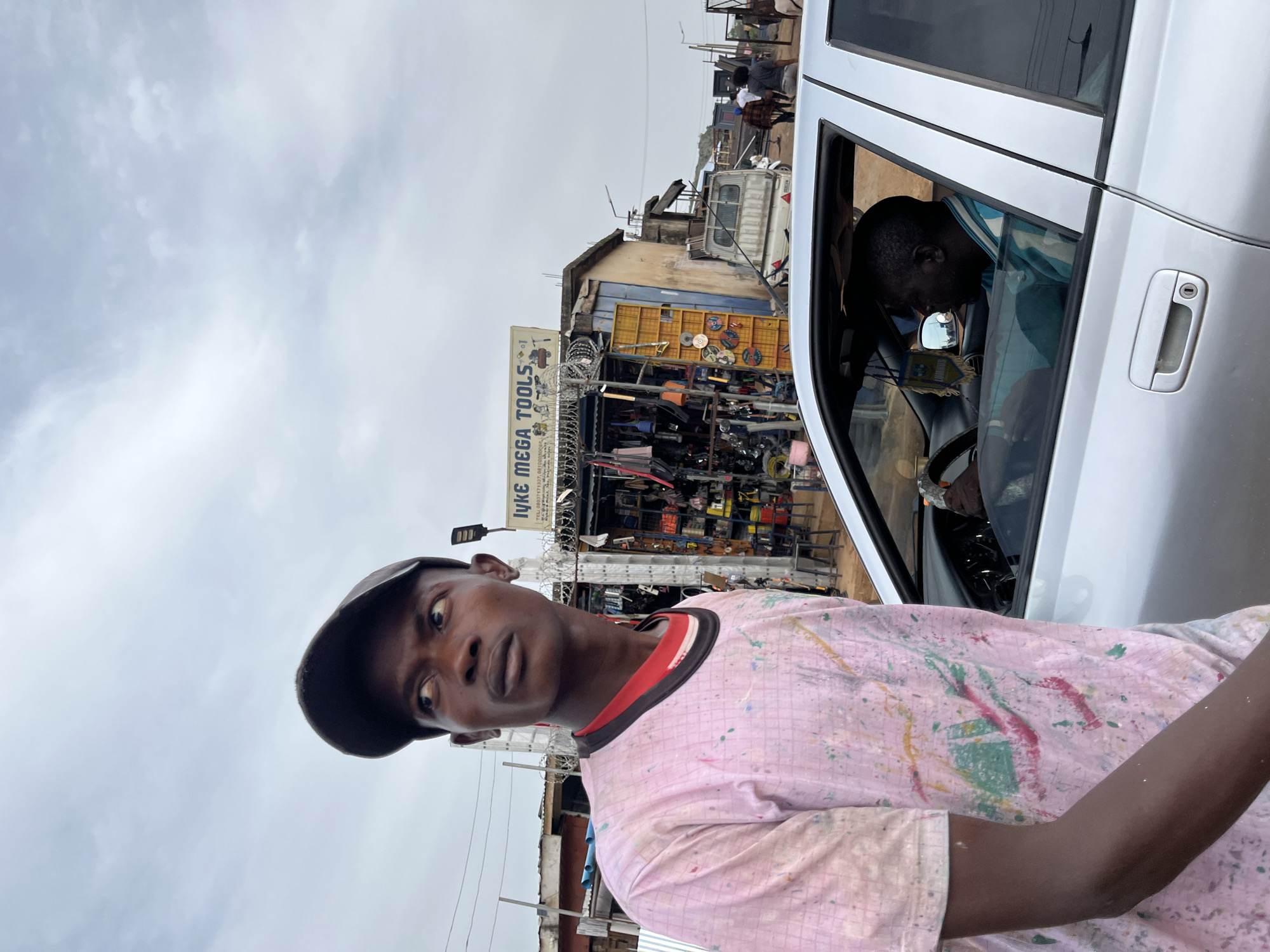 The image shows a young Black man wearing a pink t-shirt splattered with paint and a dark baseball cap. He is looking directly at the camera with a neutral expression. He is standing in front of a building that appears to be a shop with a sign that reads "IYKE MEGA TOOLS". The shop is filled with various items, including tools and mechanical parts, and is fenced with barbed wire.
To the right of the man, a silver car is parked, with its door open. A Black man is visible inside the car, looking down. The scene appears to be an outdoor market or roadside shop in Gura Top, Nigeria. The sky is overcast, suggesting it might be a cloudy day. The general atmosphere appears to be that of a busy commercial area.