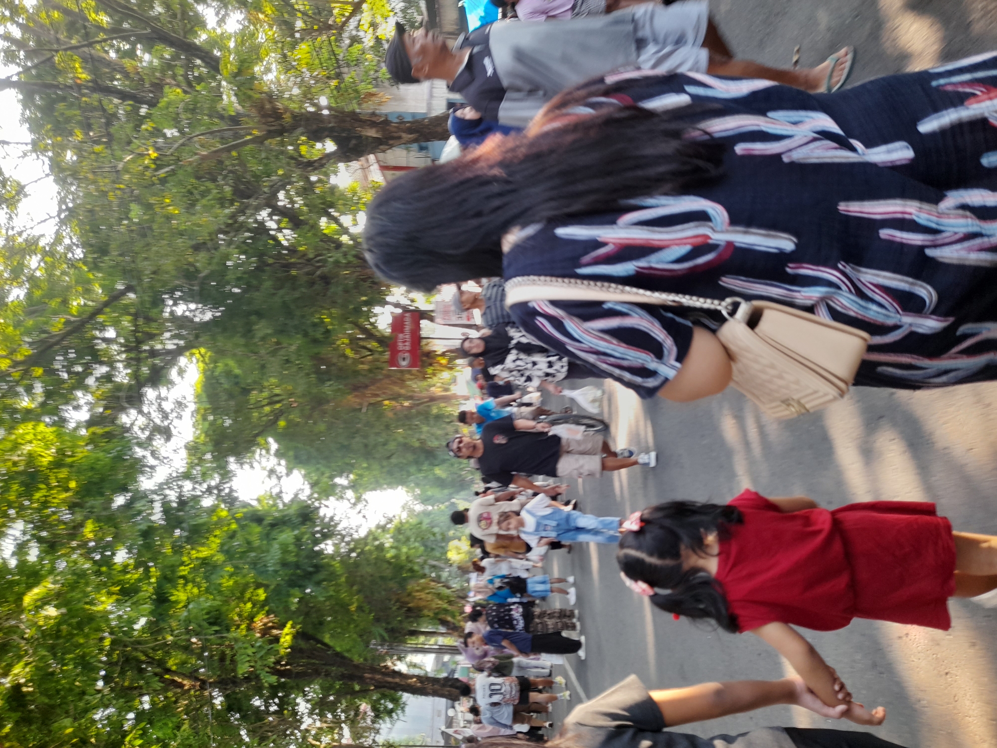A group of people are walking on a street in Purwodadi, Indonesia, lined with trees. In the foreground, a young girl in a red dress is holding hands with an adult. Behind them, a woman in a dark blue dress with a cactus pattern walks away from the camera, carrying a beige purse. In the background, other pedestrians are visible, some of whom are wearing casual clothing. The street is paved, and sunlight filters through the trees, casting shadows on the ground.