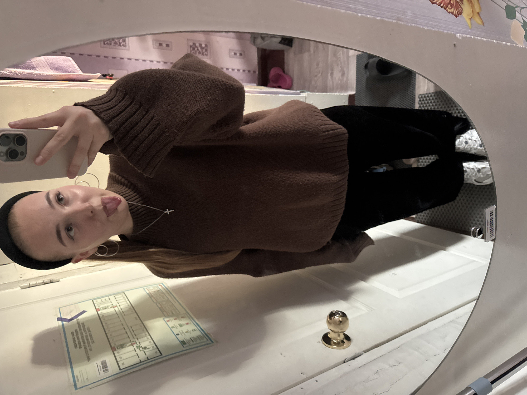 A young woman in a brown sweater, black pants, and white sneakers is taking a selfie in a mirror. She is wearing a black headband, silver hoop earrings, and a silver necklace with a cross pendant. She is sticking her tongue out. The mirror is hanging on a white door with a gold doorknob. A diagram, possibly a building plan, is affixed to the door with a piece of blue tape. A hat, slippers, and other objects are visible behind the woman in the mirror's reflection. The environment appears to be inside an apartment or residence. The city is not discernible from the image.