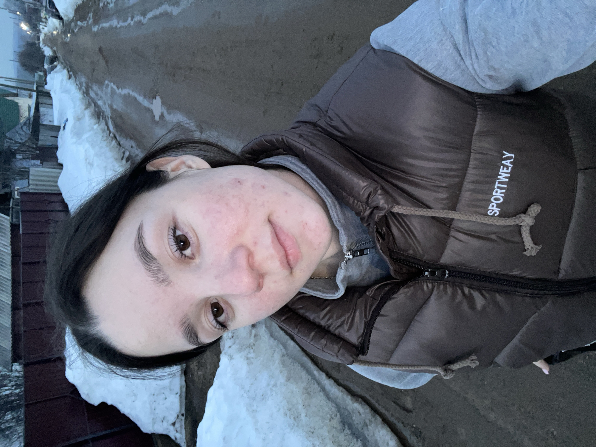 A young female adult, appearing to be in her late teens or early twenties, poses for a selfie on an unpaved road in Ольховка, Russia. She has dark hair, brown eyes, and fair skin displaying visible facial redness and minor blemishes. Her expression is neutral, and she looks directly at the camera. She wears a dark brown, possibly bronze, quilted puffer vest over a light grey hooded sweatshirt with a zipper; the vest is branded with "SPORTWEAY" in white text on the left chest. A thin silver chain is visible around her neck. The road beneath her is a muddy, unpaved surface with visible tire tracks and puddles from melting snow. Large, irregularly shaped snowbanks line both sides of the road, indicating a late winter or early spring environment. In the background, properties are separated from the road by fences, including a dark brown corrugated metal fence on the left. Distant residential structures and utility poles are visible under an overcast, dim sky, suggesting twilight or low light conditions. No other individuals are present.