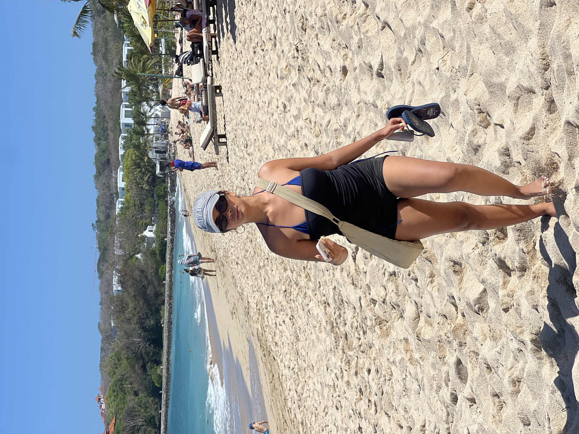 A woman is walking on a sandy beach in Kuta Selatan, Indonesia, towards the camera. She is wearing a black one-piece swimsuit with a blue bikini top underneath, black shorts, and a grey baseball cap. She has sunglasses on and is carrying a beige shoulder bag and a pair of blue flip-flops. The beach is wide and sandy, with gentle waves rolling in from the turquoise ocean. In the background, palm trees and resort buildings are visible, as well as other beachgoers relaxing on lounge chairs and walking along the shore. The sky is clear and blue.